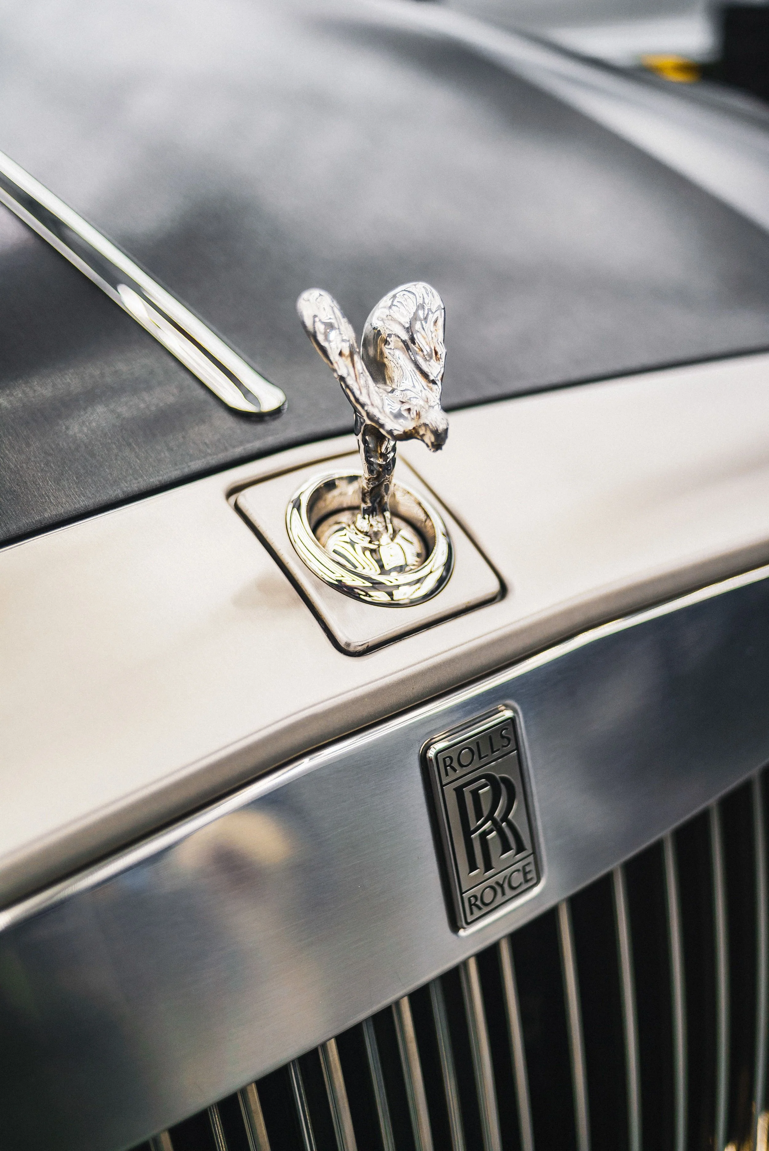 Close-up of a vintage Rolls-Royce car hood ornament, featuring the Spirit of Ecstasy figure, with a part of the car's grille and emblem visible below.