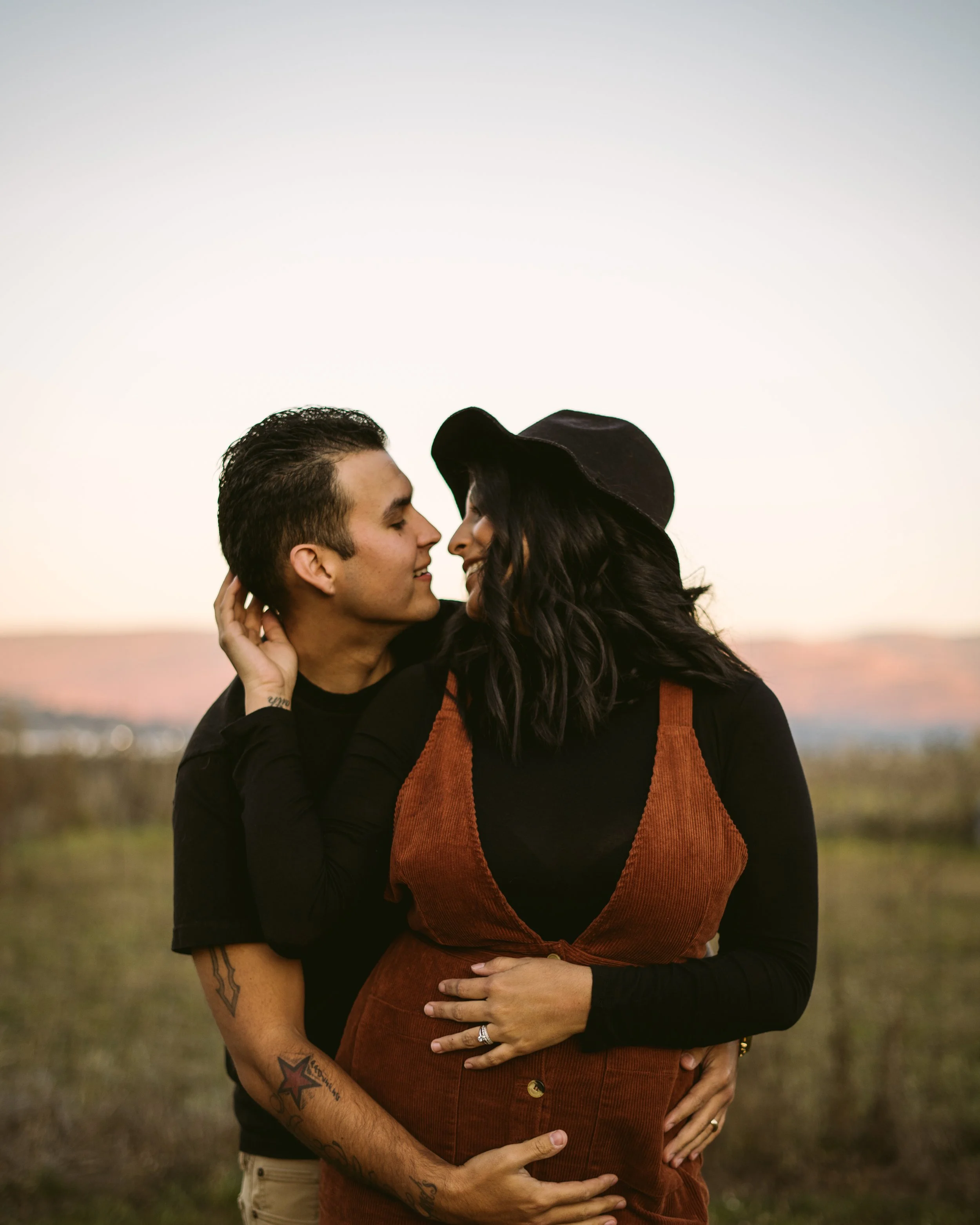 A couple standing outdoors at sunset, embracing, with the man behind the woman, both with their eyes closed, smiling gently, in a natural setting with a blurred landscape in the background.
