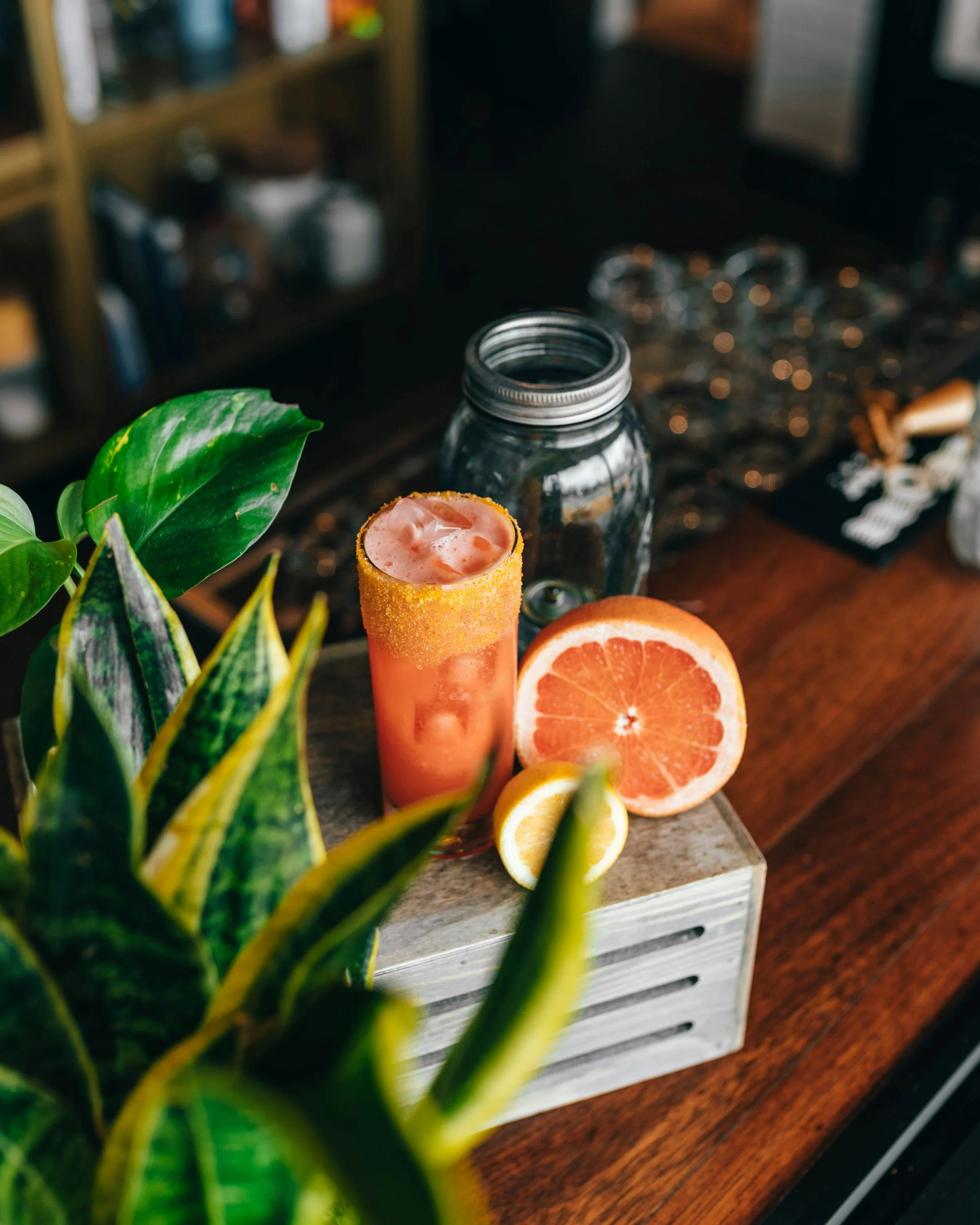 Close-up of a fresh grapefruit half on a wooden surface, sliced lemon slices, a tall glass filled with pink grapefruit juice, and a plant with green leaves in the foreground, with a blurred bar or kitchen in the background.