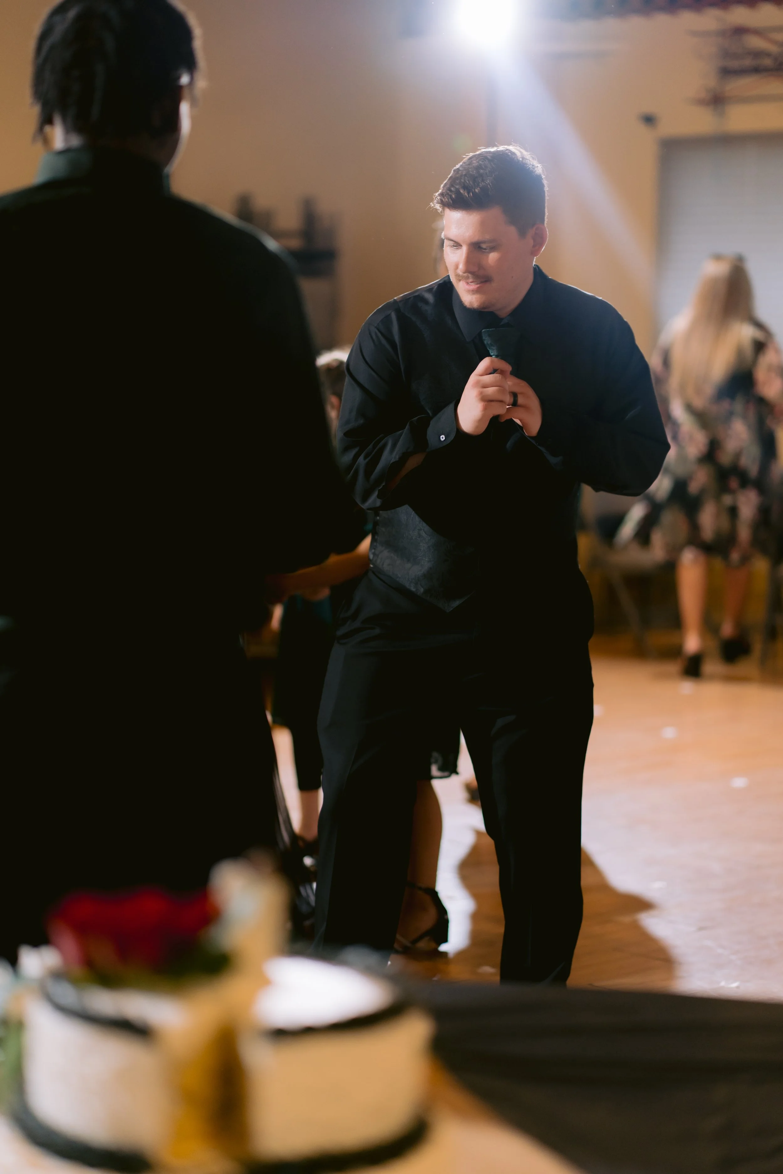 A man in a black shirt and vest dancing with a smile, looking down, in a decorated event space. Blurred people and a woman in a floral dress are in the background, with a dessert table in the foreground.