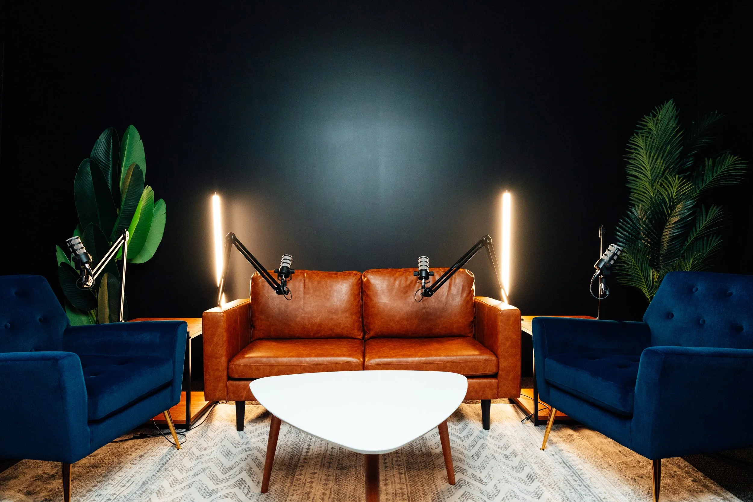 A modern podcast studio with a brown leather sofa, two blue velvet armchairs, a white coffee table, and two tall floor lamps against a dark wall. There are green leafy plants on both sides of the setup.