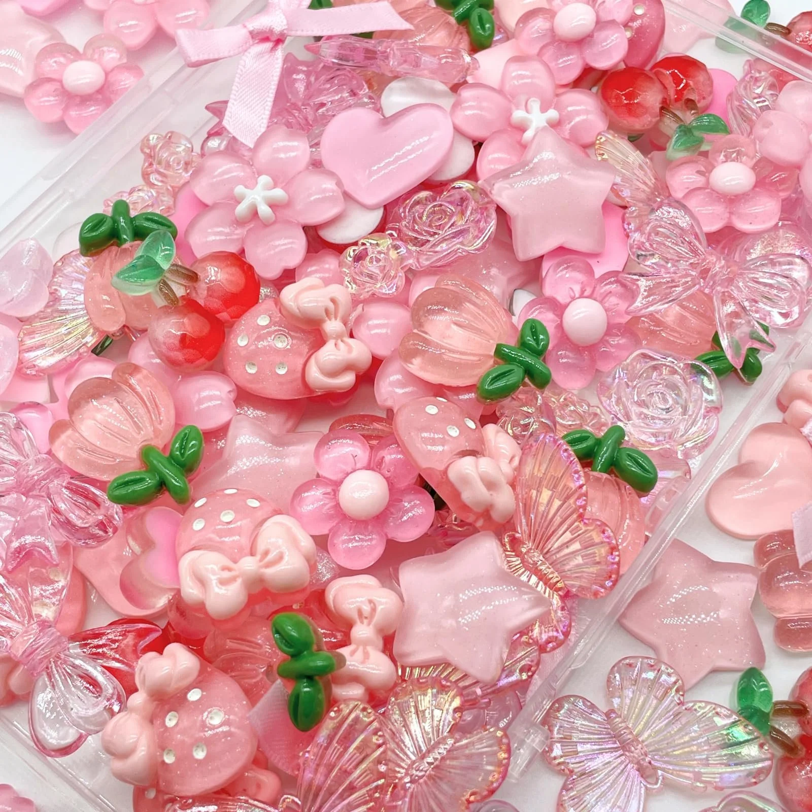 Assorted pink, red, and green glass and plastic jewelry charms and embellishments scattered across a surface, including hearts, stars, roses, butterflies, cherries, bows, and flowers.