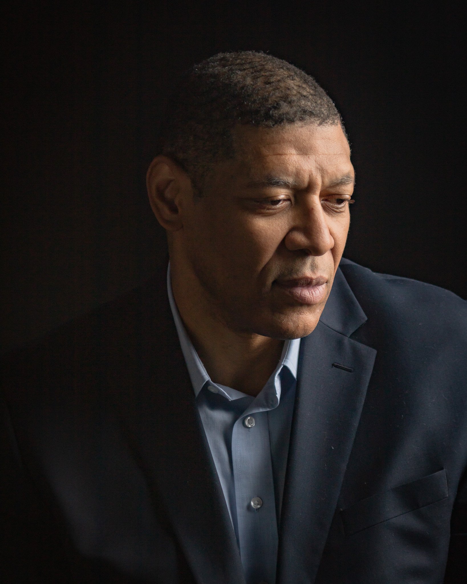 Close-up of a man with dark skin, wearing a navy blue blazer and light blue shirt, looking downward with a serious expression. The background is black.