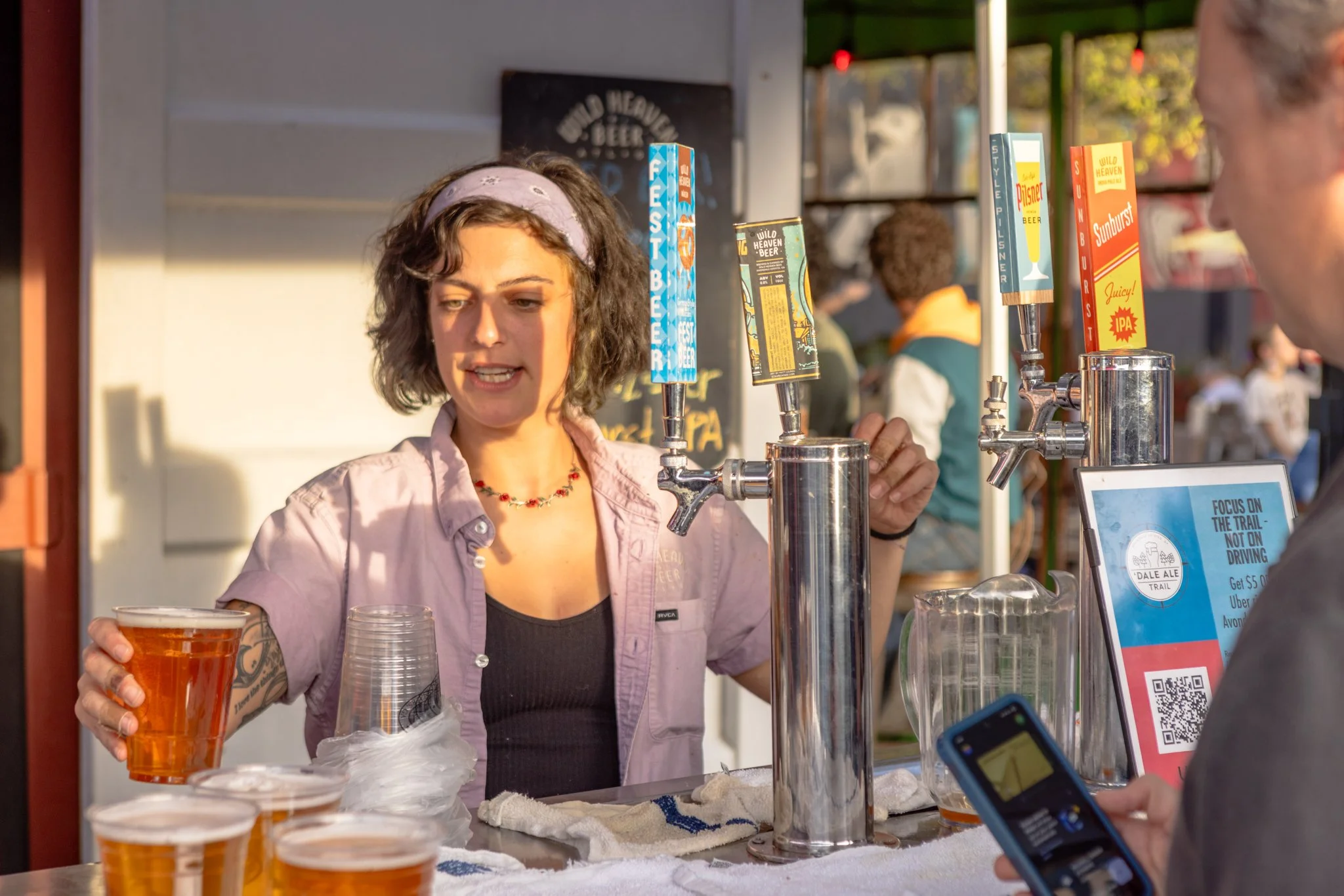 A woman with curly hair, wearing a pink shirt and a headband, is pouring beer from a tap at a beverage stand, with cups filled with beer on the counter in front of her.