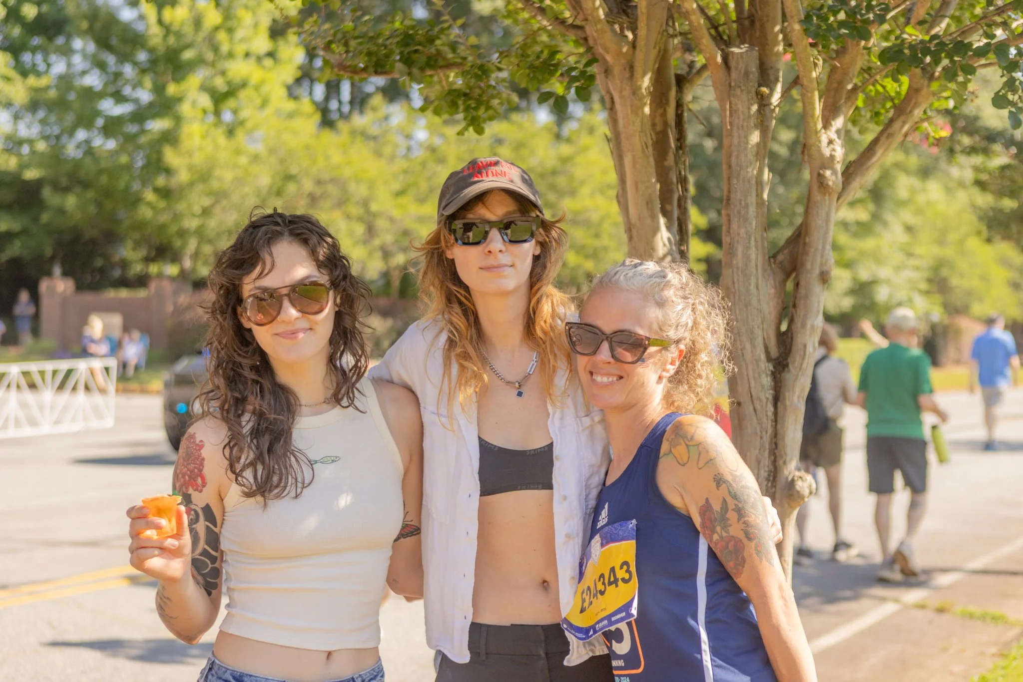 Three women are standing outdoors at a park, smiling and posing for the photo with a tree and other people in the background. One woman is holding a small object, possibly a snack.