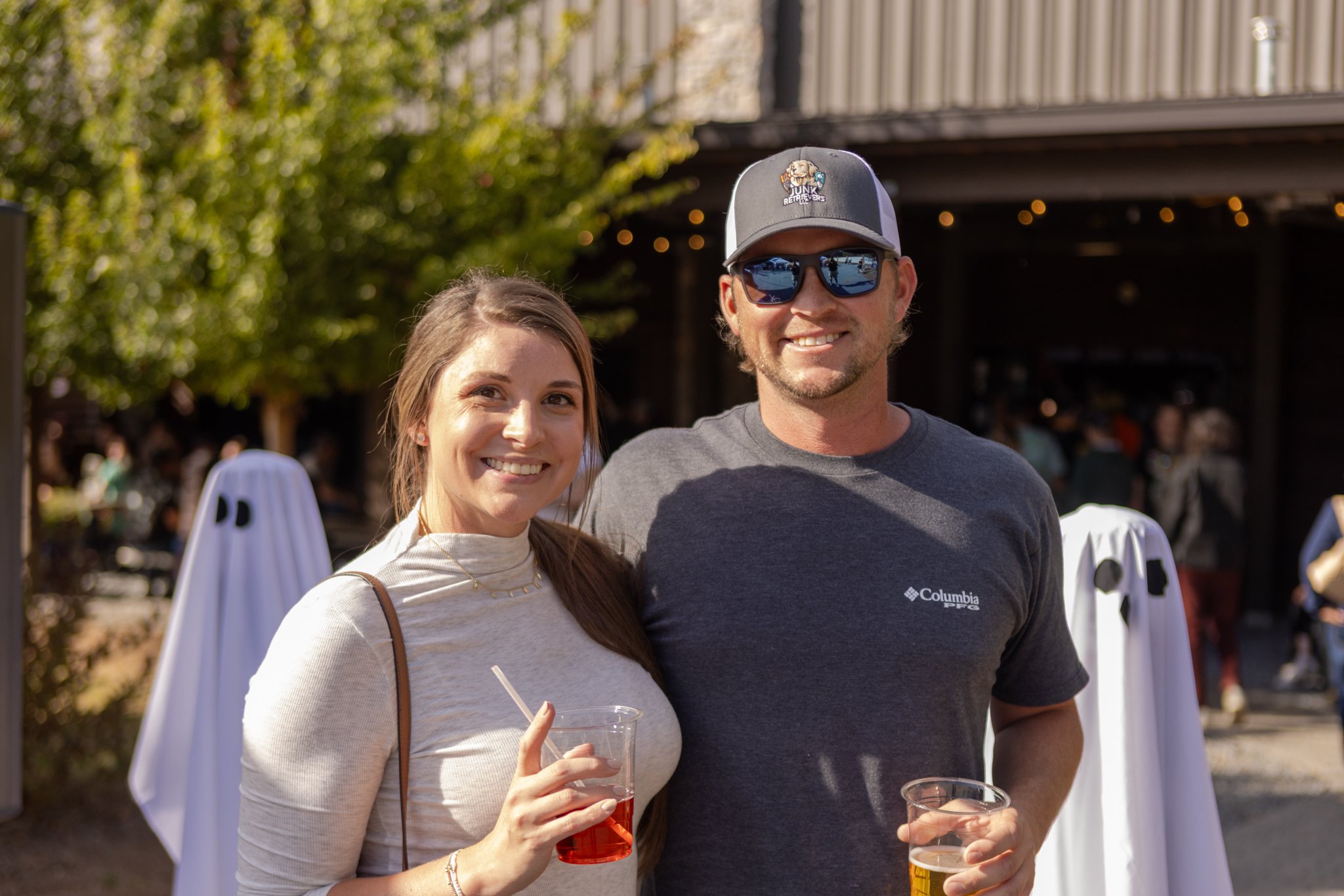 A smiling woman with long brown hair, wearing a beige top and holding a drink, stands next to a smiling man with short hair, wearing sunglasses, a gray cap, and a gray t-shirt, also holding a drink. They are outdoors at a social event with blurred tr
