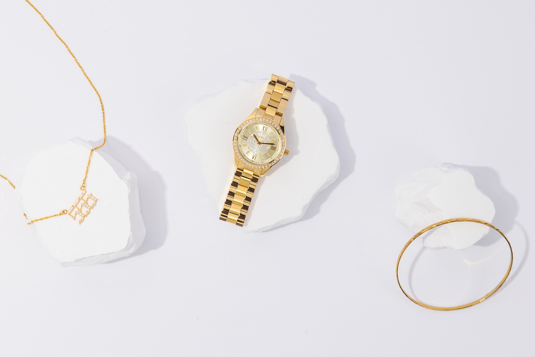 A gold watch with a diamond-encrusted face, surrounded by gold jewelry including a necklace, bracelet, and earrings, displayed on white rocks against a light grey background.