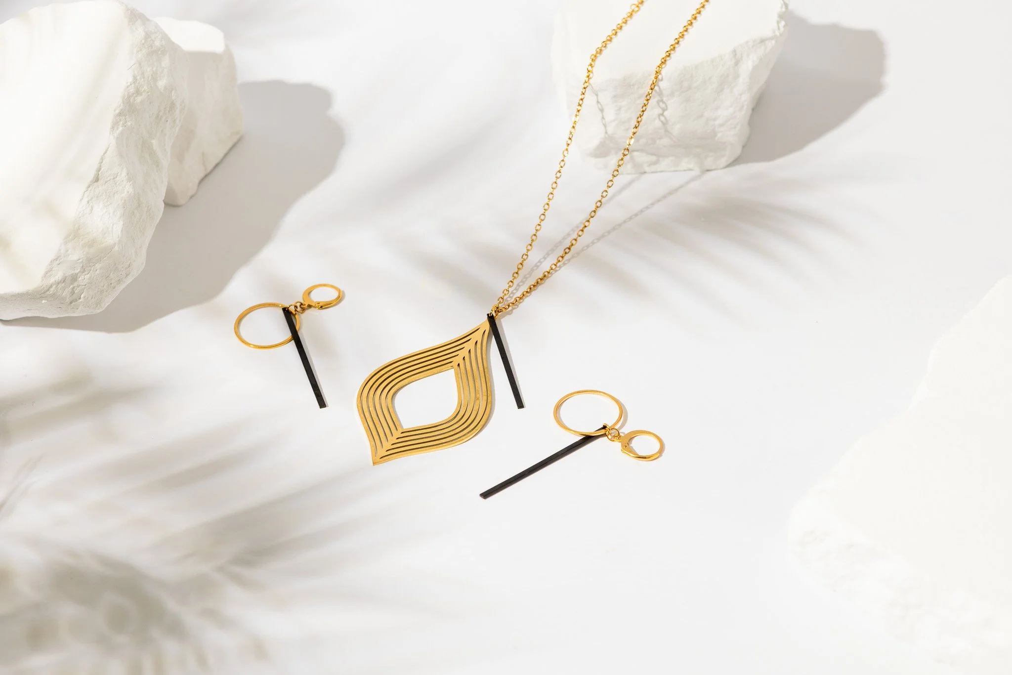 Gold and black jewelry set including a necklace with a gold abstract fan-shaped pendant, matching earrings with gold hoops and black accents, and a gold chain, displayed on white textured rocks and paper.