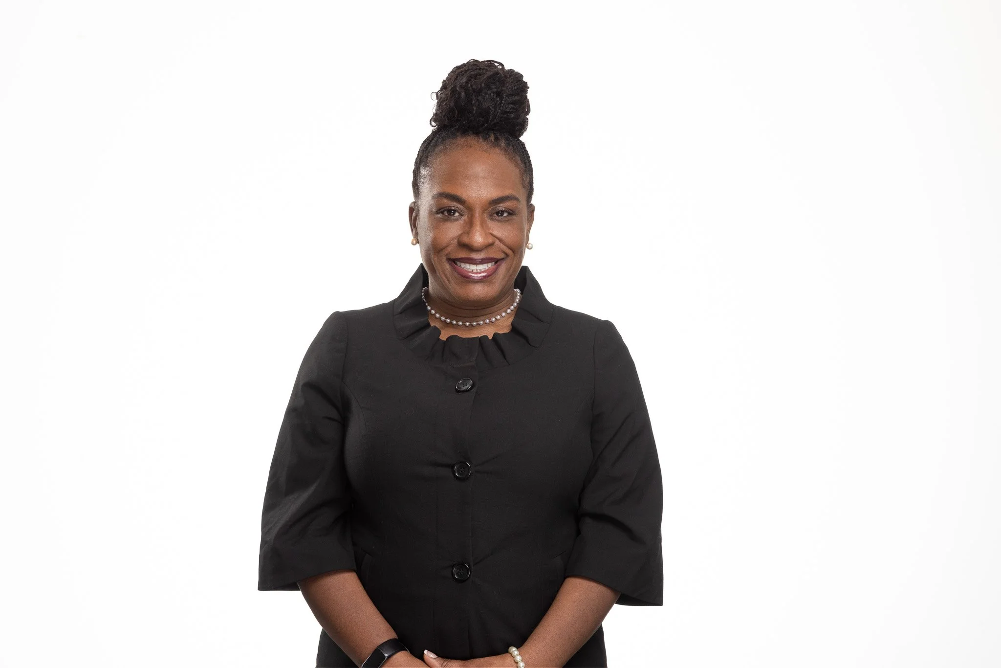 Portrait of a smiling African American woman in a black suit, wearing pearl earrings and a pearl necklace, with her hair styled in a high bun, standing against a white background.