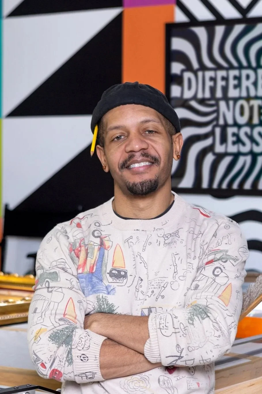 A man with a beard, wearing a black cap, earrings, and a white sweatshirt with abstract drawings, standing with arms crossed in front of a colorful, geometric, and artistic mural background.