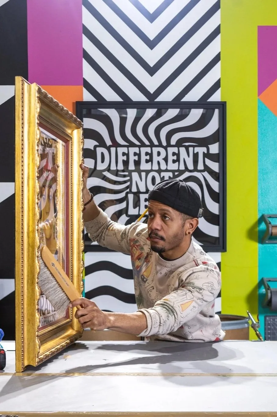 A man cleaning a framed painting with a gold frame in an art studio with colorful abstract walls and a black-and-white wavy background art piece that reads 'DIFFERENT NOT LESS'.