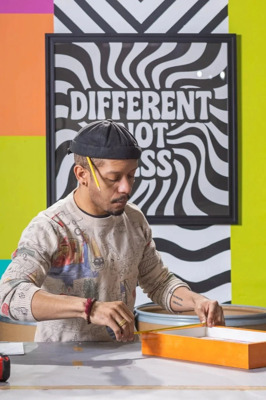 A man with a beard, wearing a black cap and glasses, is measuring a box with a yellow ruler. He has tattoos on his arms and is wearing a beige sweater with colorful sketches. The background features a black and white swirling pattern with the words "