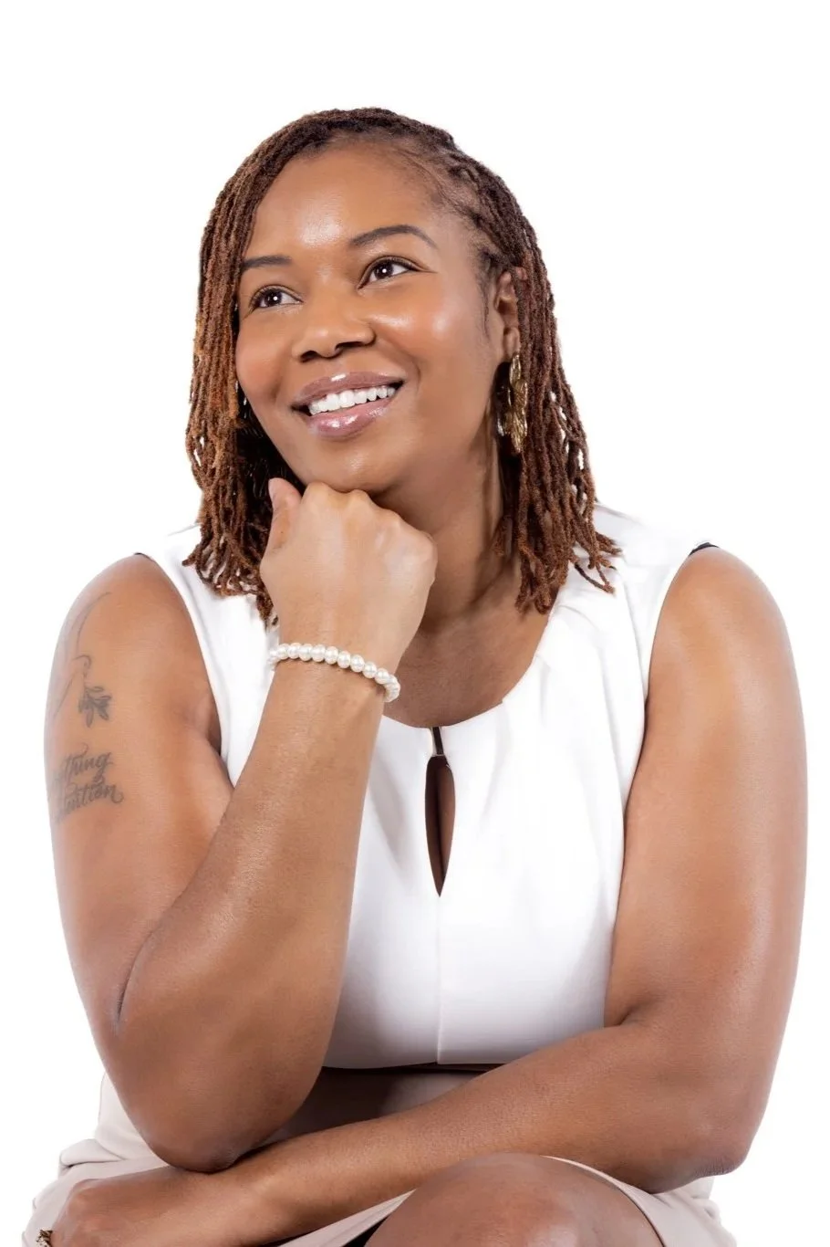 A woman with shoulder-length dreadlocks wearing a white sleeveless top, smiling, with her chin resting on her hand, against a plain white background.