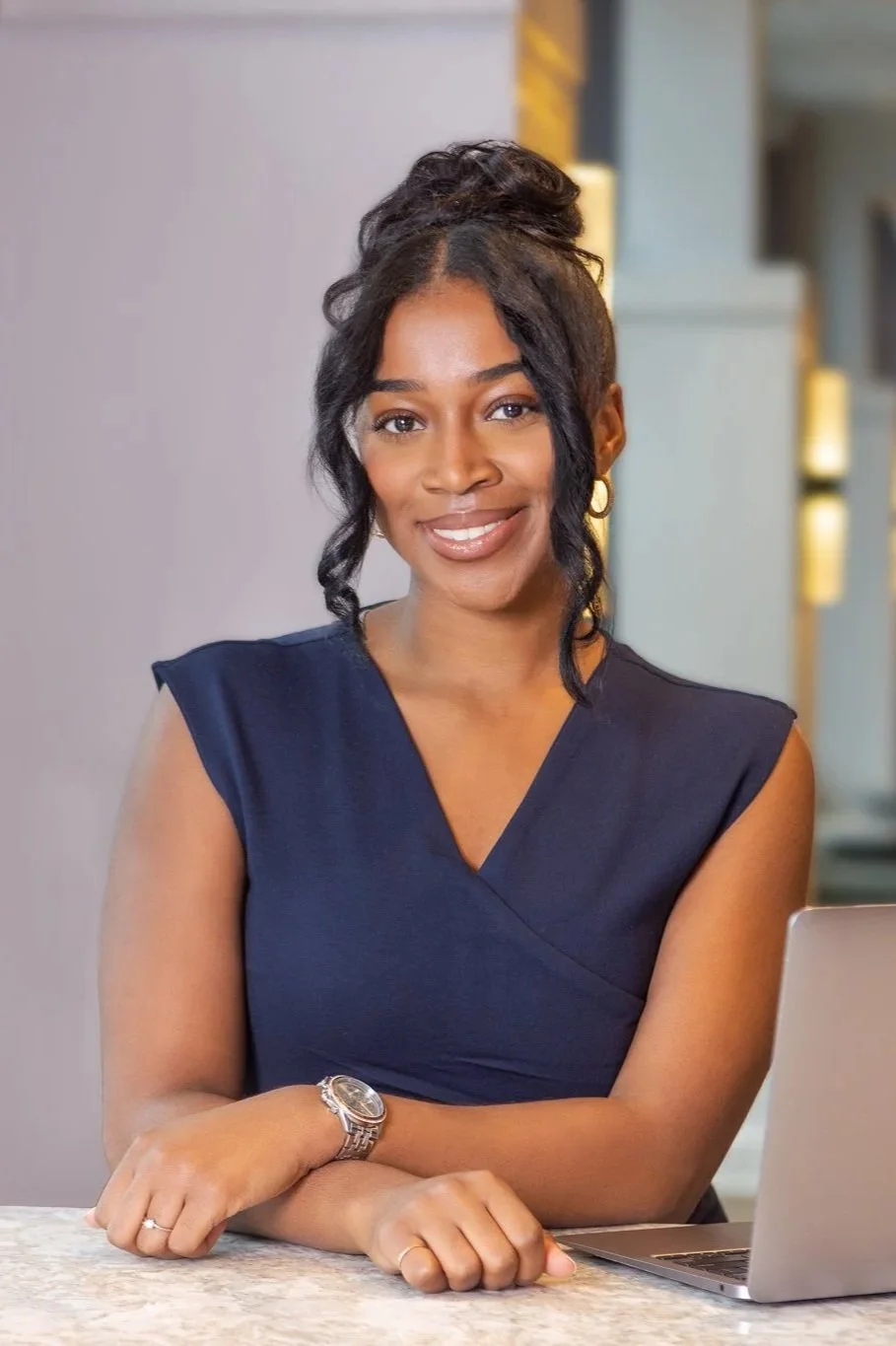 A woman sitting at a desk with a laptop, smiling at the camera, wearing a dark sleeveless top, with her arms crossed and a silver watch on her left wrist.