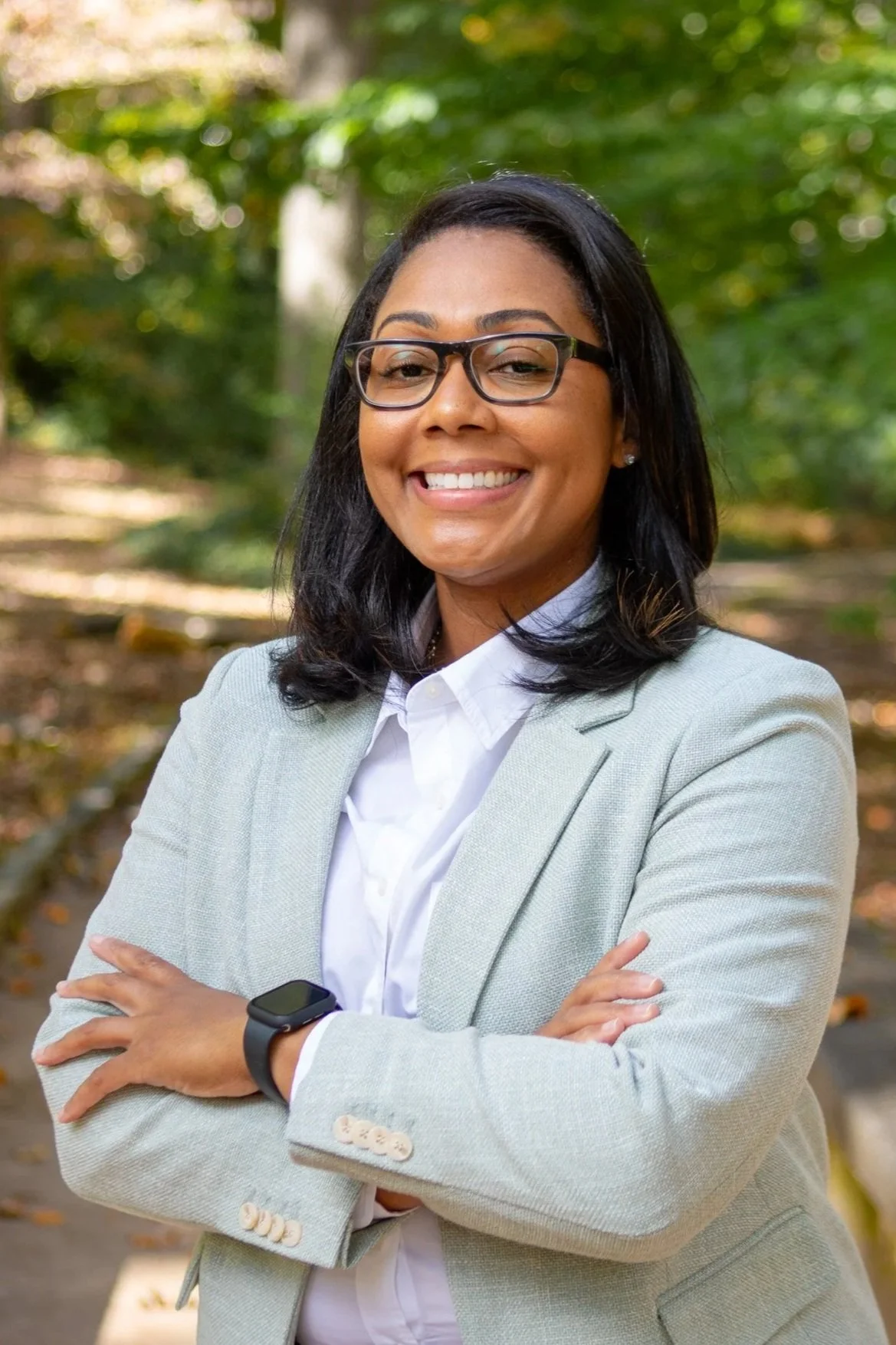 A professional woman smiling outdoors in a park, wearing glasses, a light gray blazer, white shirt, and a smartwatch, with arms crossed.