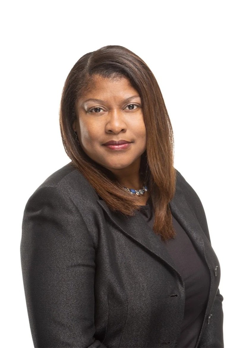 A professional woman with brown hair wearing a dark blazer and a necklace, posing against a white background.
