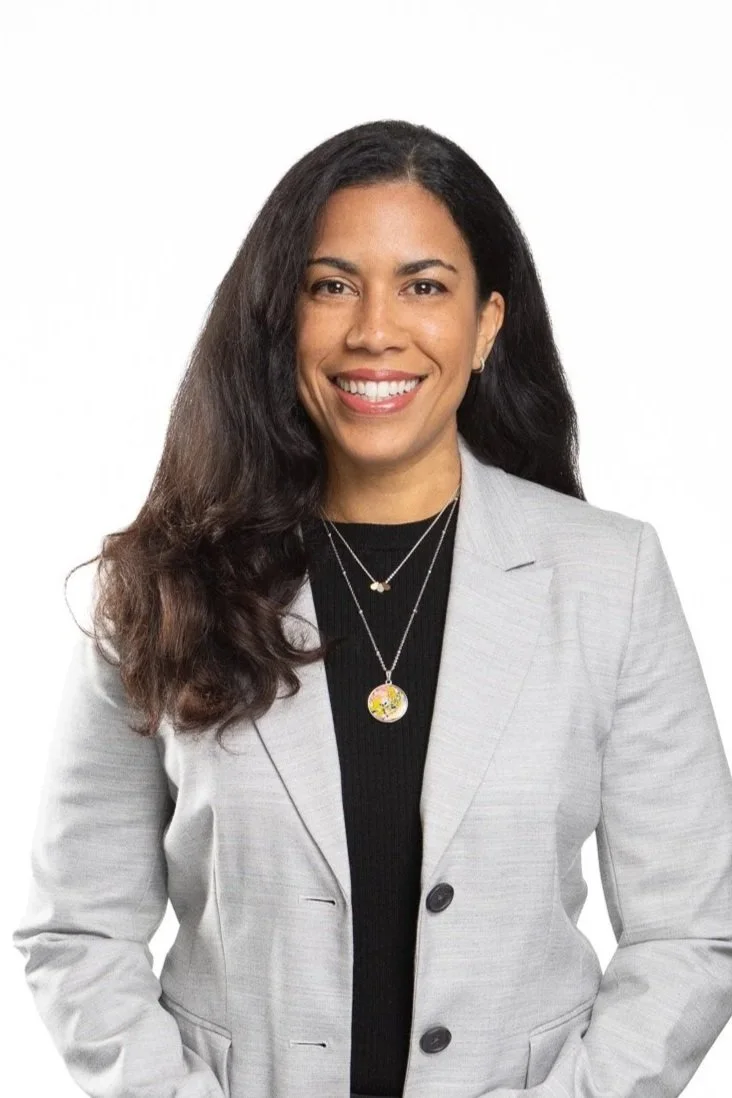 Portrait of a woman with long dark hair, wearing a gray blazer over a black top, and smiling with white teeth, against a white background.
