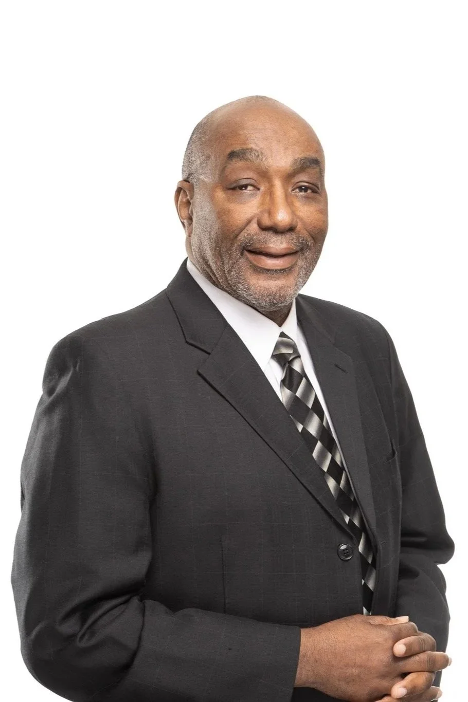 An older man in a black suit with a white shirt and black and white striped tie, smiling slightly, standing against a plain white background.