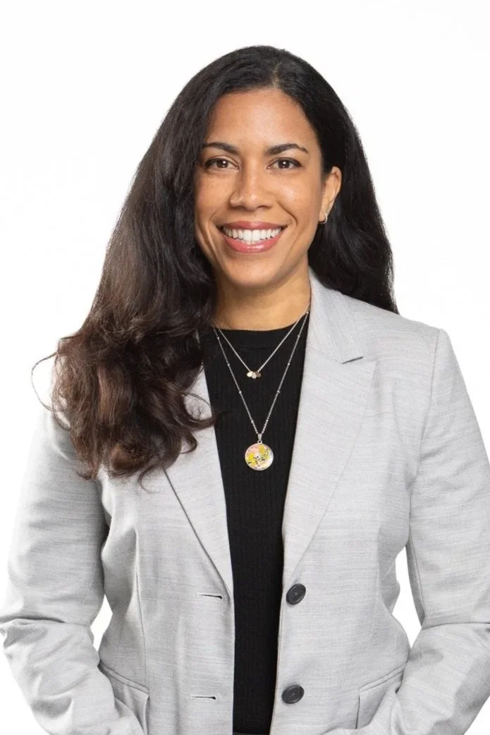 A woman with long dark hair smiling, wearing a light gray blazer over a black top and layered necklaces, against a plain white background.