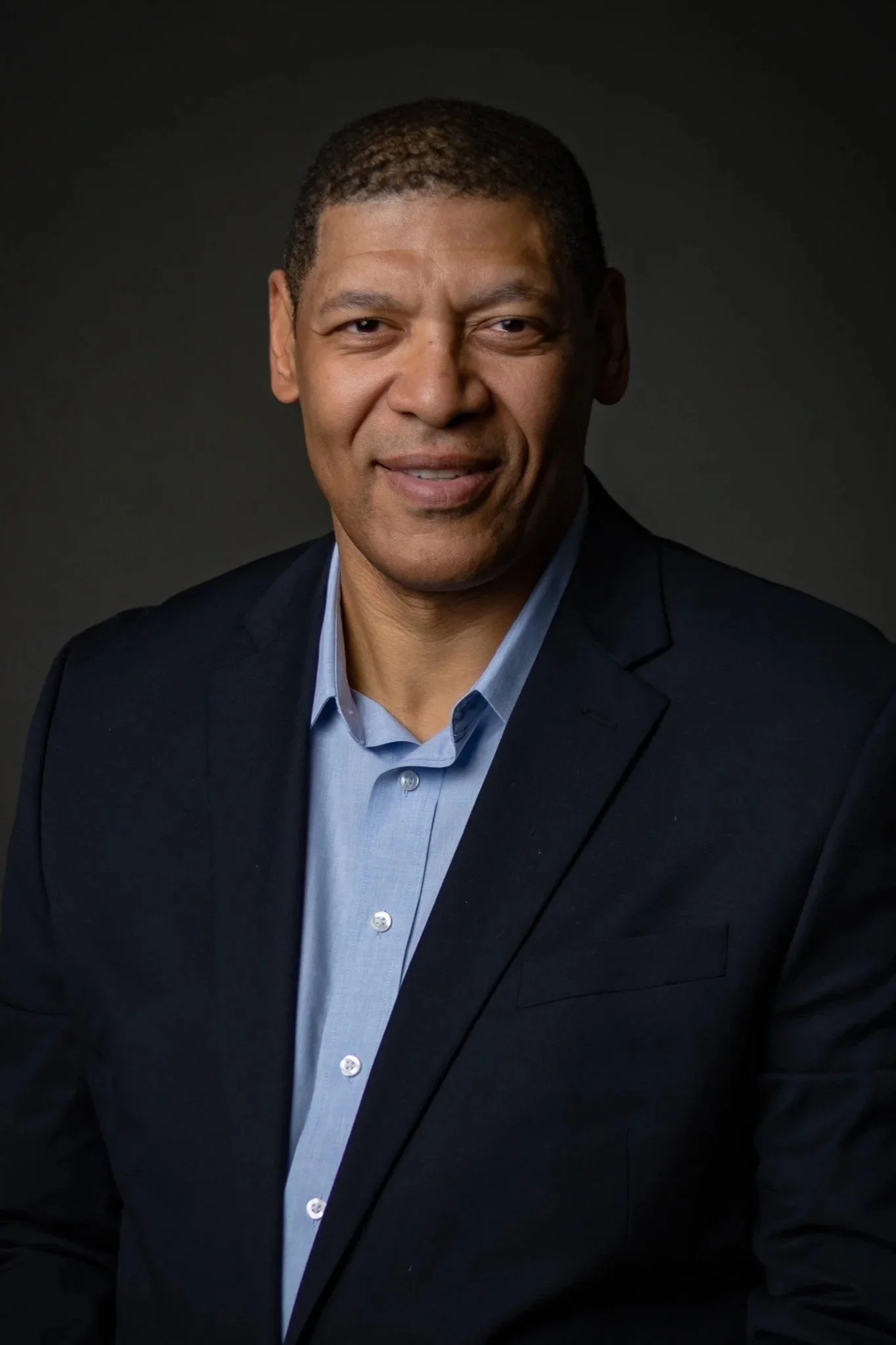 Portrait of a middle-aged man with short curly hair, wearing a dark navy suit jacket and a light blue dress shirt, smiling slightly against a dark background.