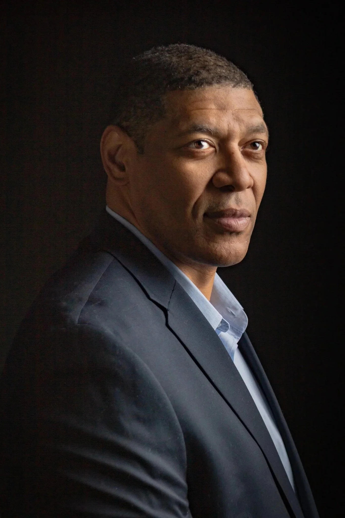 A man in a business suit looking over his shoulder, against a dark background.