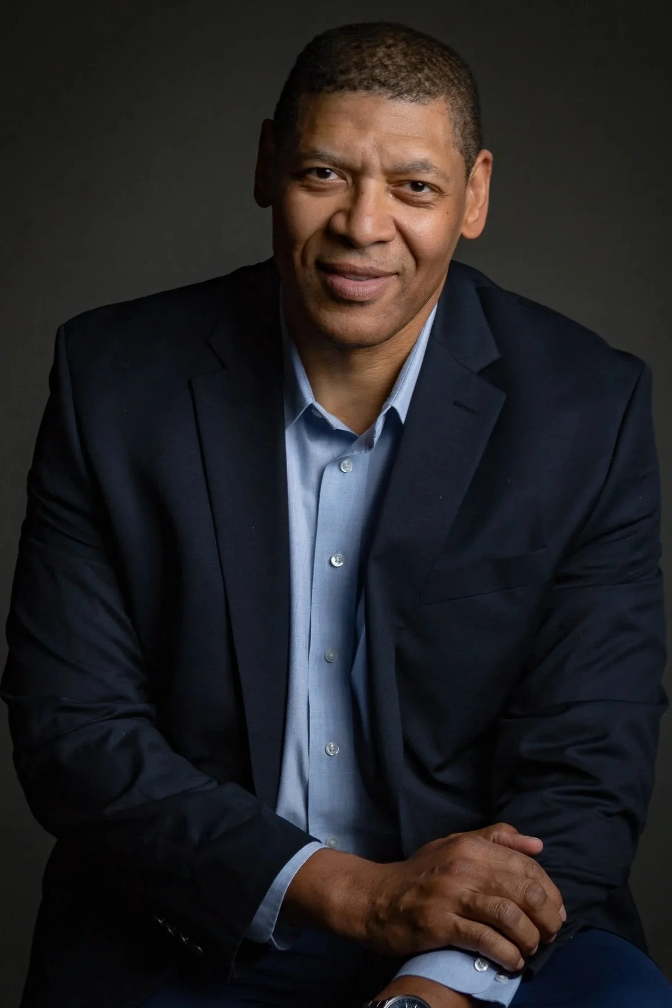 Portrait of a man in a dark suit and light blue shirt, sitting with hands clasped in front of him against a dark background.