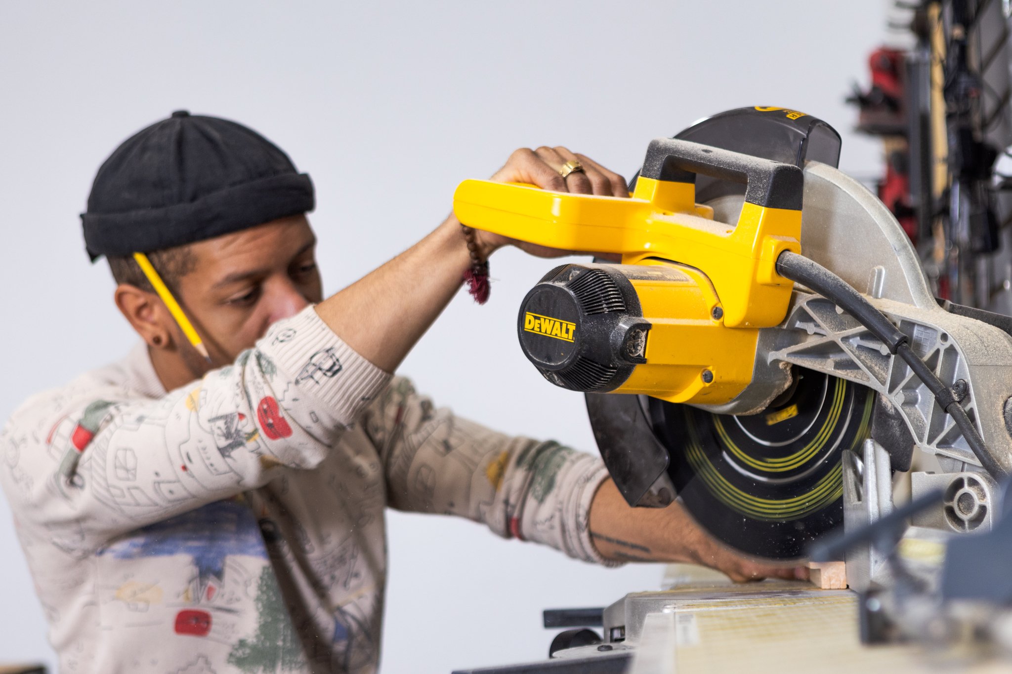 A person wearing a black beanie, safety glasses, and a paint-splattered shirt using a yellow DeWalt miter saw to cut a piece of wood.