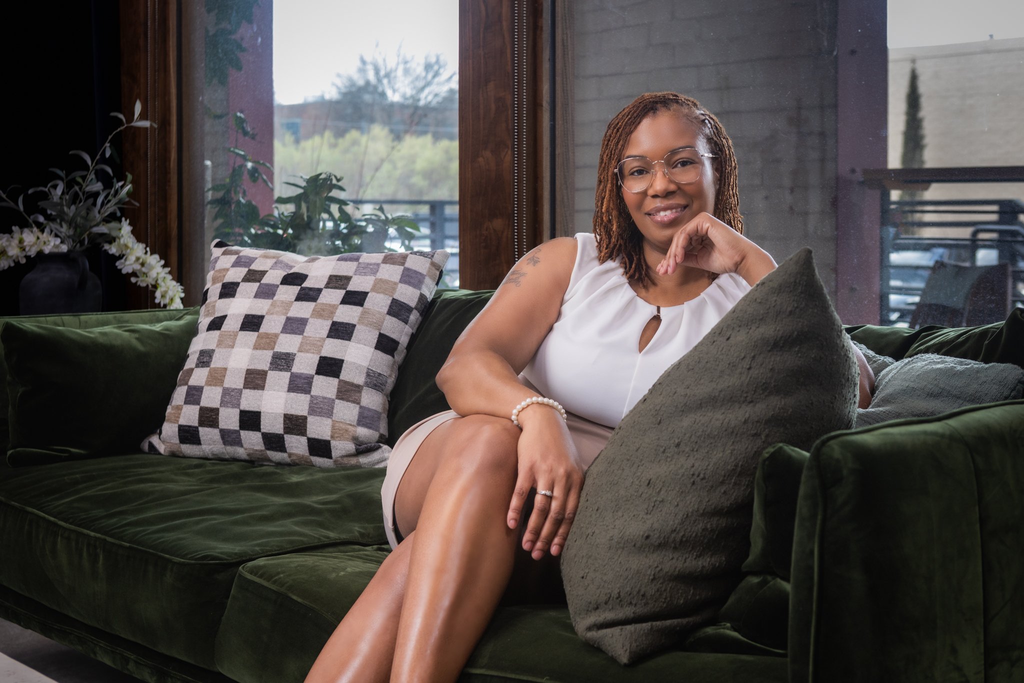A woman with glasses and dreadlocks sitting on a green velvet couch near a window, smiling and resting her chin on her hand, surrounded by pillows and plants.