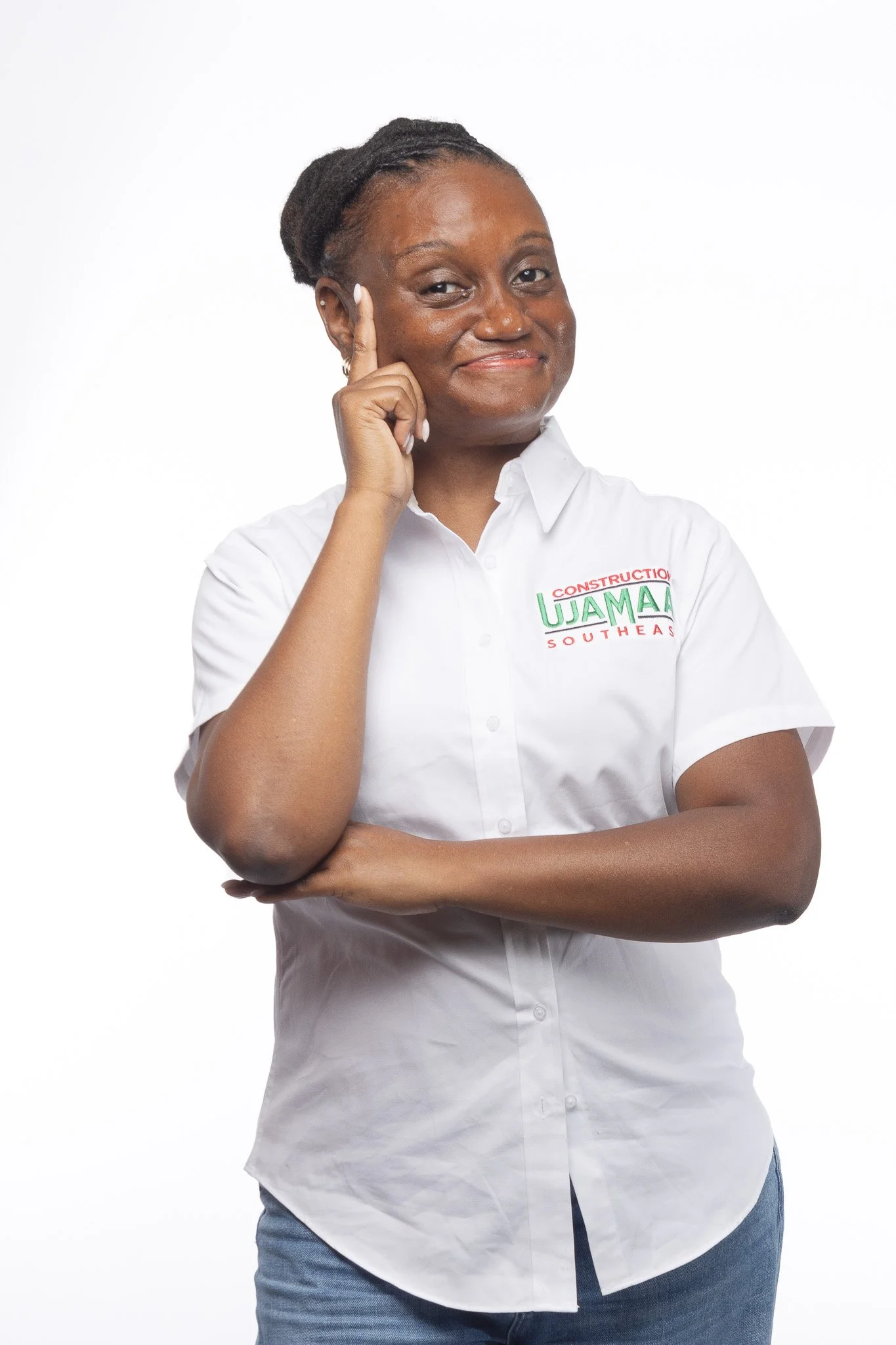 A woman posing with her hand on her head and smiling; she is wearing a white shirt with a logo and blue jeans, against a plain white background.