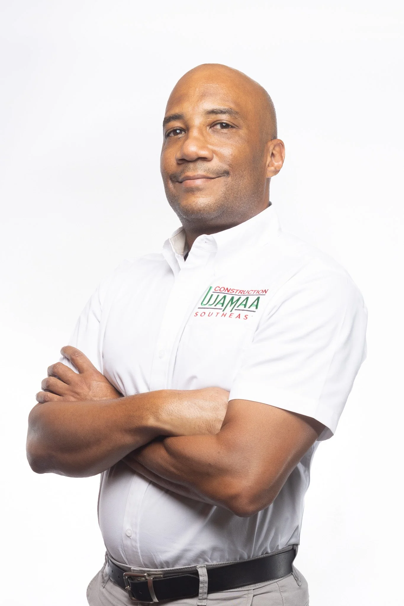 A man with a bald head and medium skin tone, wearing a white shirt with a logo that reads "Construction UJAMAA SouthEast," standing with arms crossed against a plain white background.