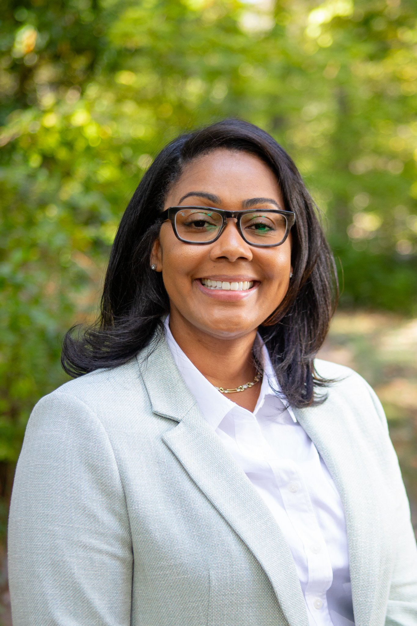 A woman with black hair, wearing glasses, a white shirt, and a light-colored blazer, smiling outdoors with blurred green trees in the background.