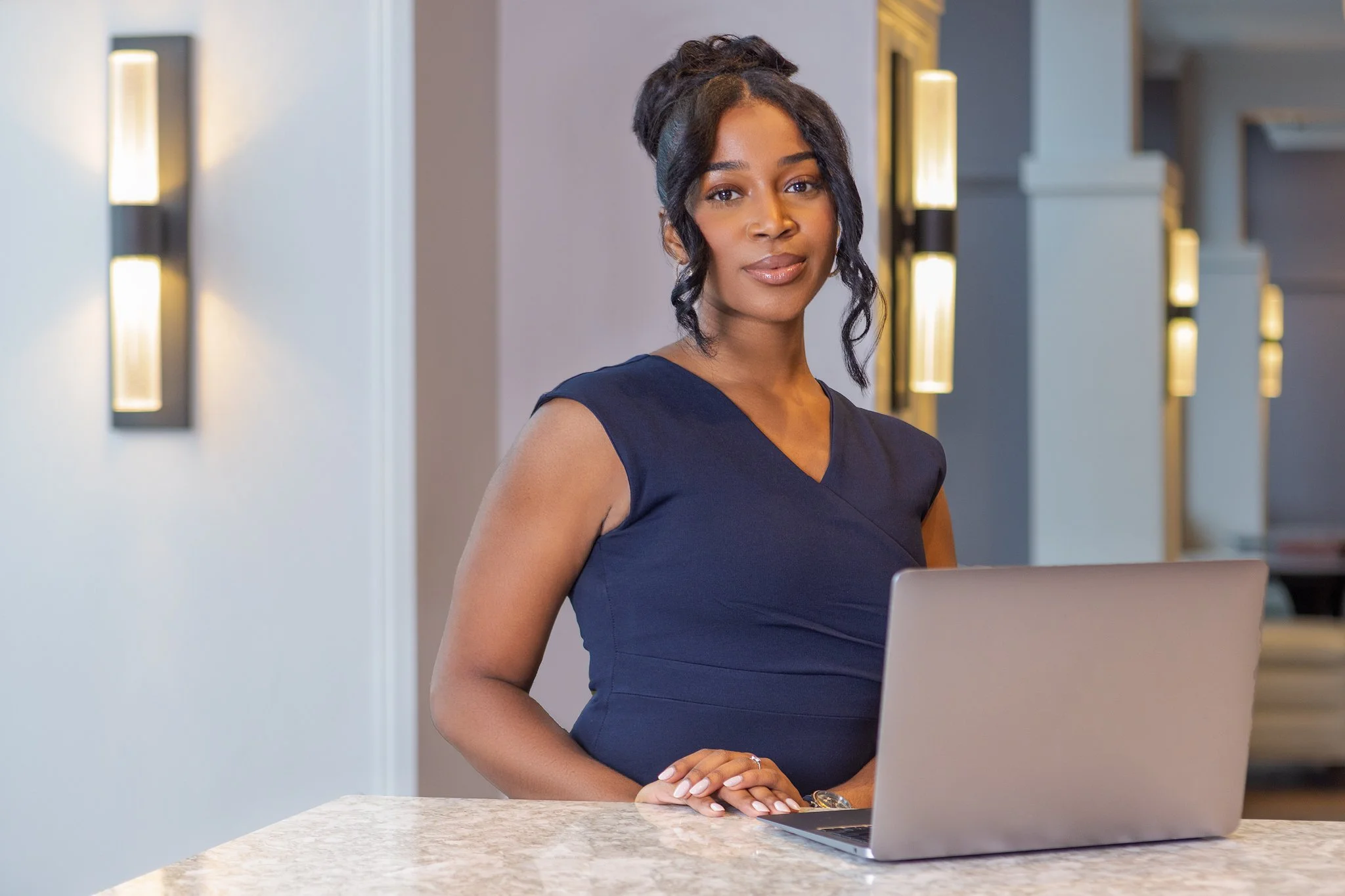 A woman with dark hair styled in waves, wearing a navy blue sleeveless top, standing behind a marble kitchen counter with a laptop in front of her, in a modern well-lit interior.