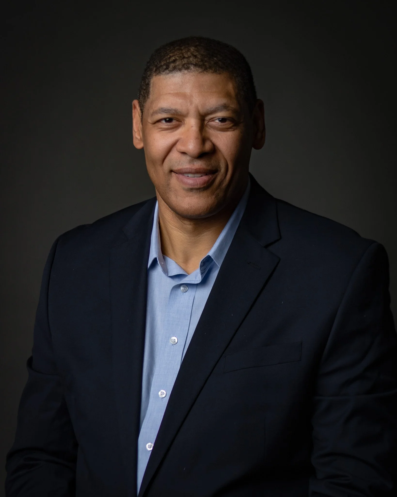 A professional portrait of a man with short dark hair, wearing a dark blazer over a light blue collared shirt, against a dark background.