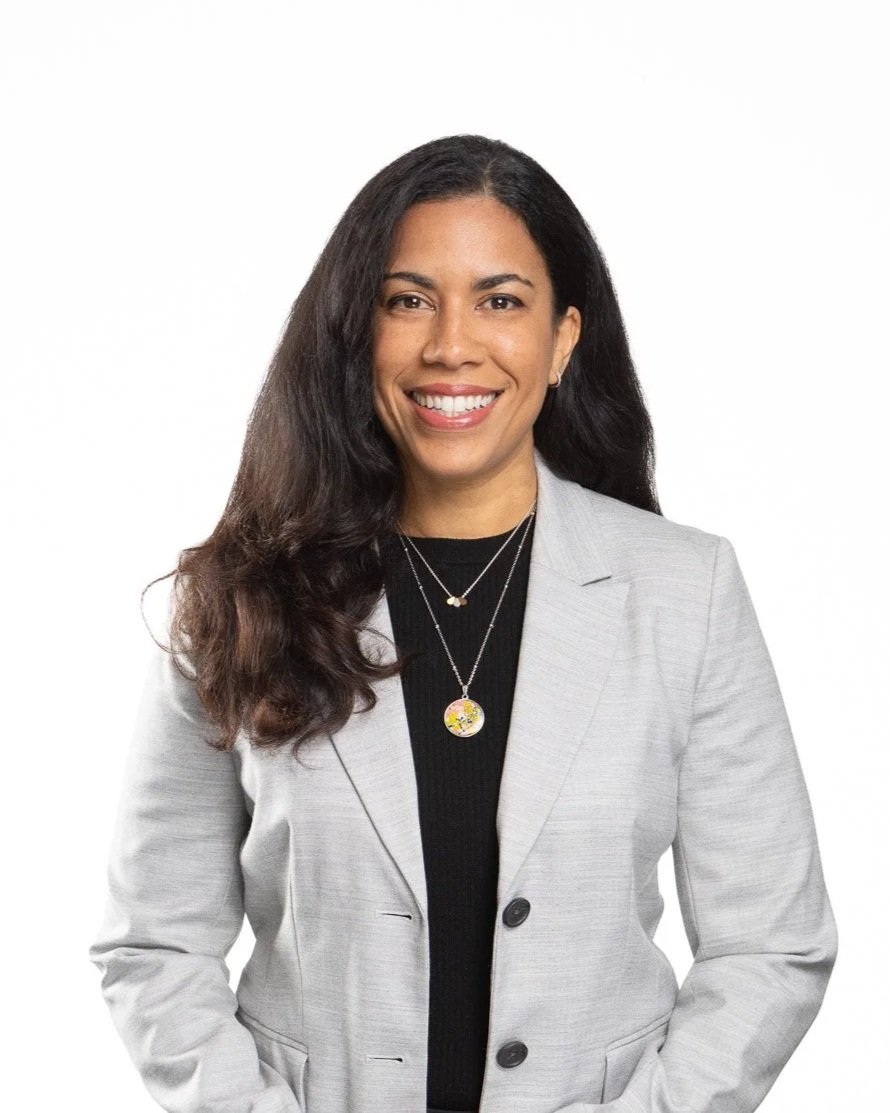 A woman with long dark hair wearing a light gray blazer over a black top, accessorized with layered necklaces, smiling against a plain white background.