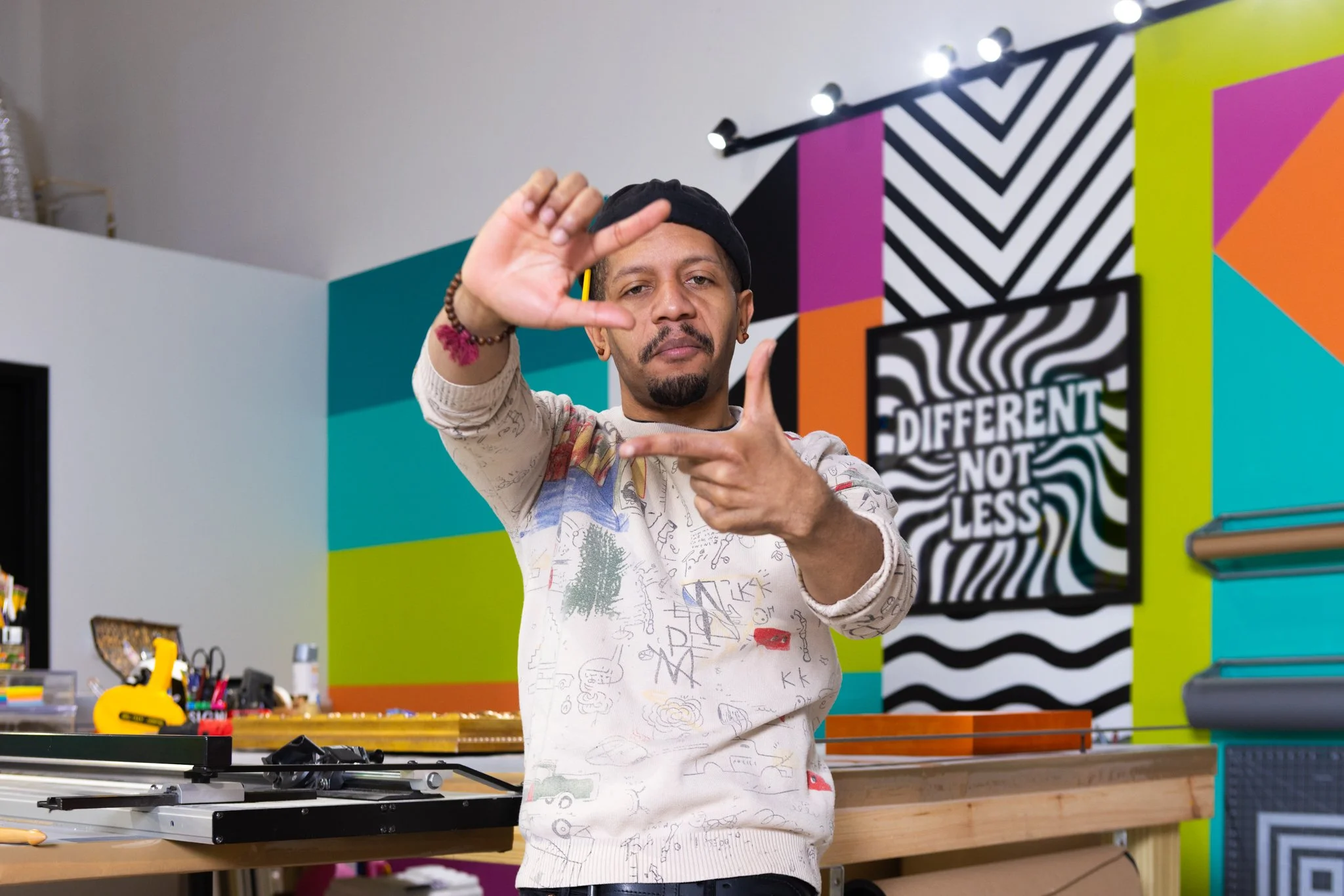 A man wearing a black cap and a white sweater making a framing gesture with his hands in a colorful art studio with striped and geometric artwork on the wall.
