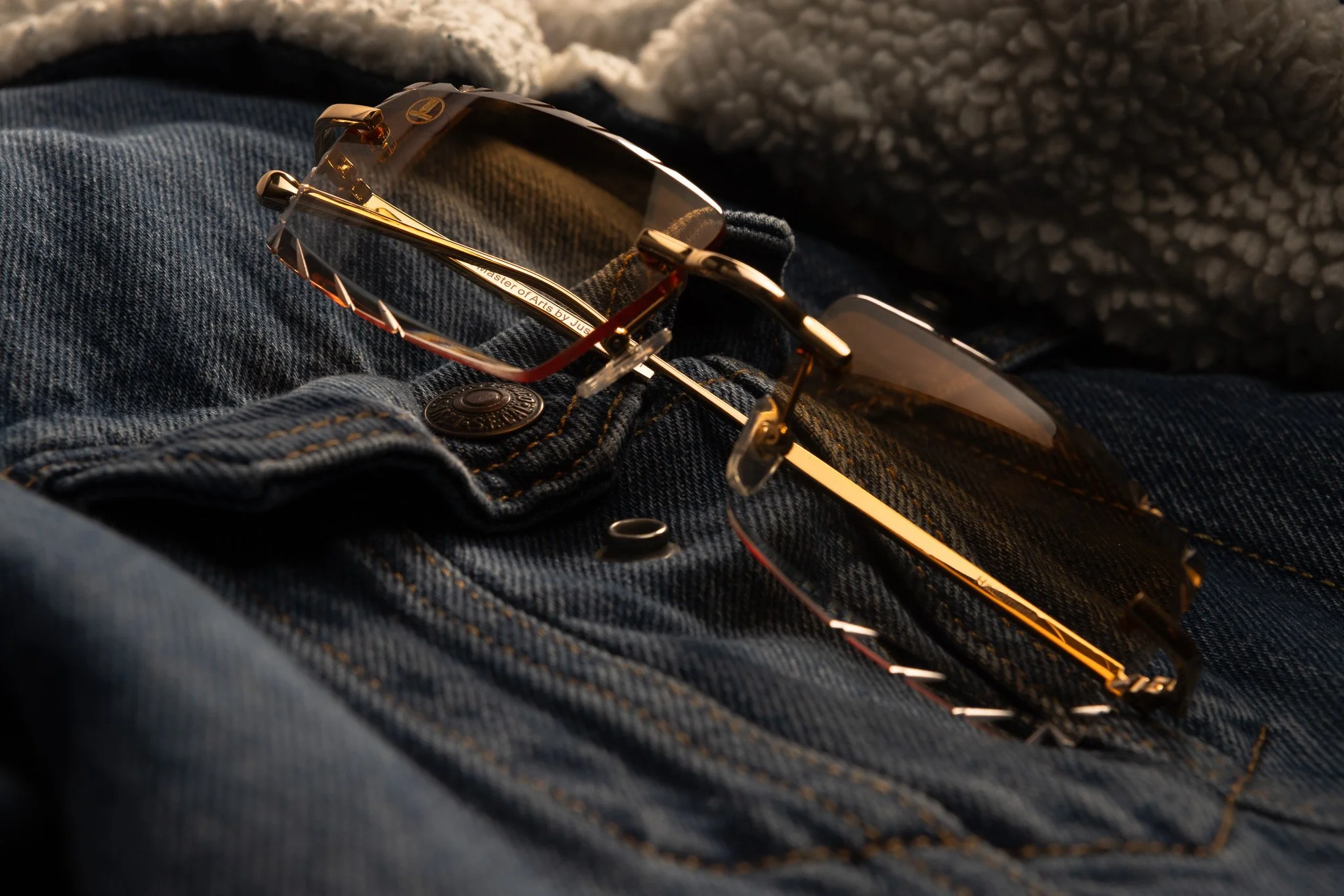 Close-up of a pair of sunglasses resting on dark denim jeans with a metal button, with a gray textured blanket or clothing item in the background.