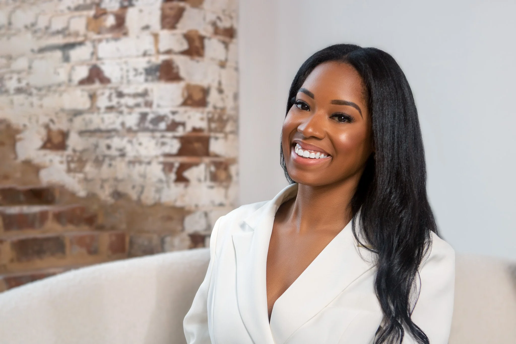 A woman with long, dark hair wearing a white blazer, smiling, in a room with a brick wall on one side and a plain wall on the other.