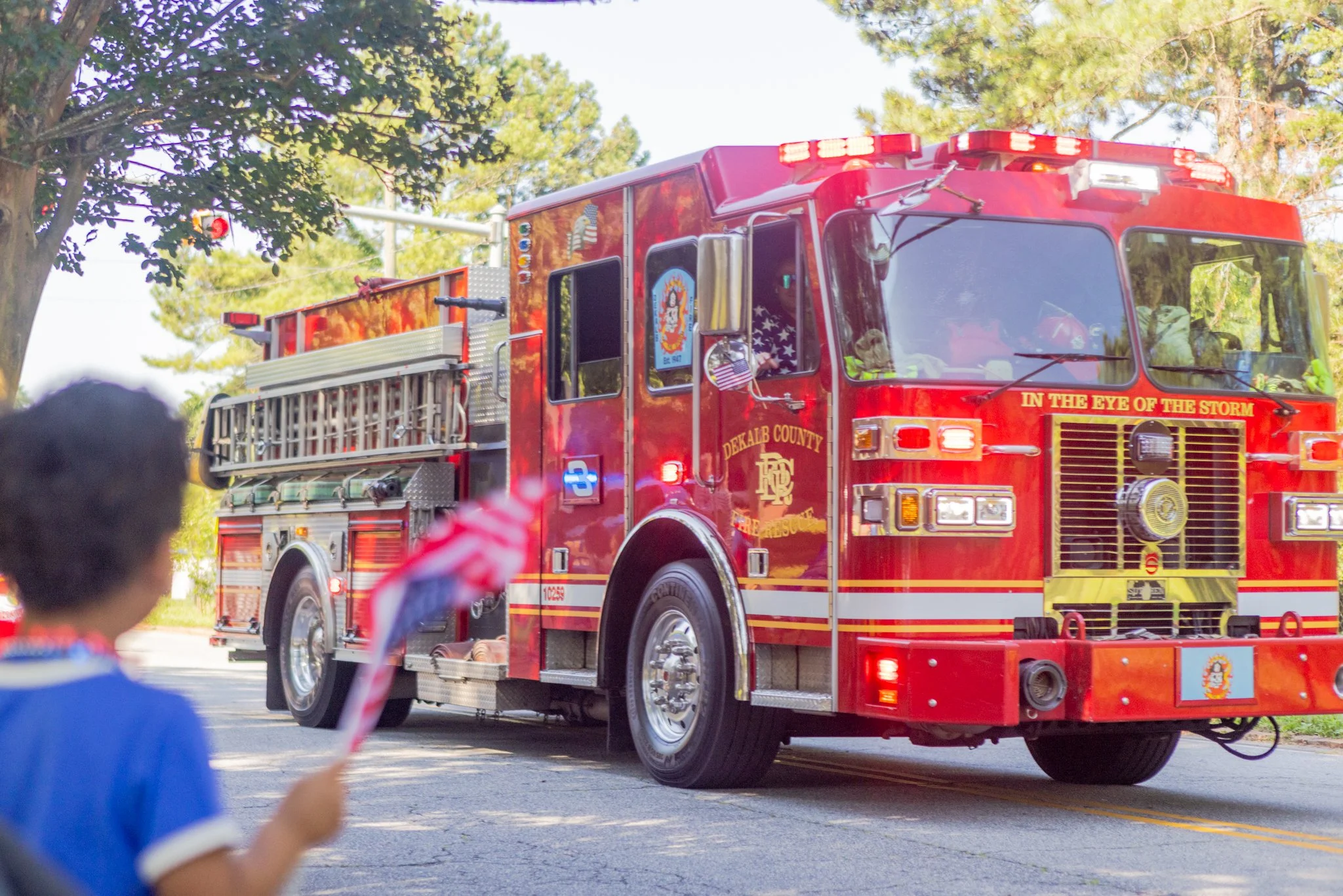 A red fire truck with the words 'In the eye of the storm' and 'DeKalb County' on the front, moving down a street lined with trees, with children watching and waving in the foreground.