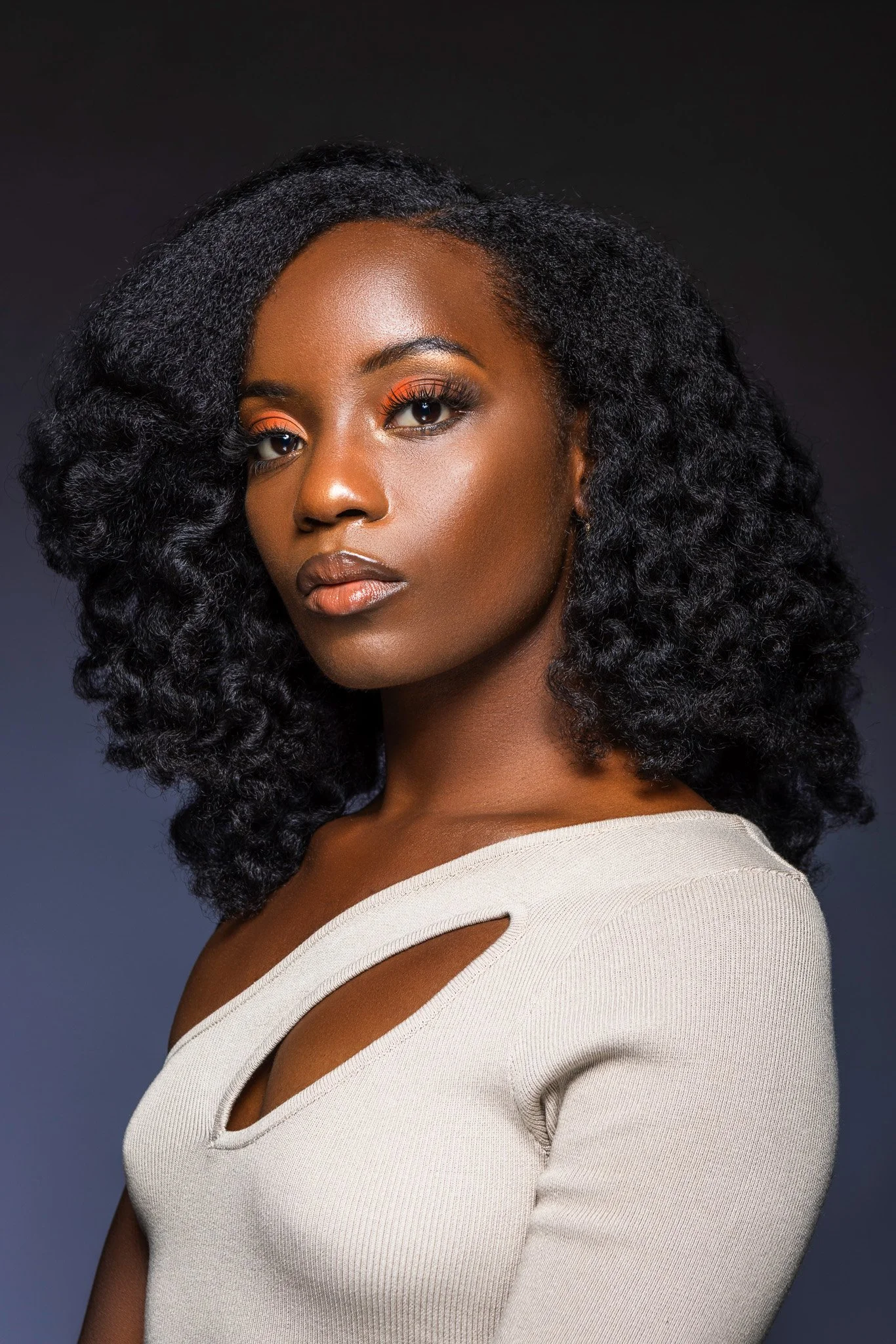 A woman with dark curly hair and brown skin wearing orange eyeshadow and a beige top with cutout details on the shoulders.