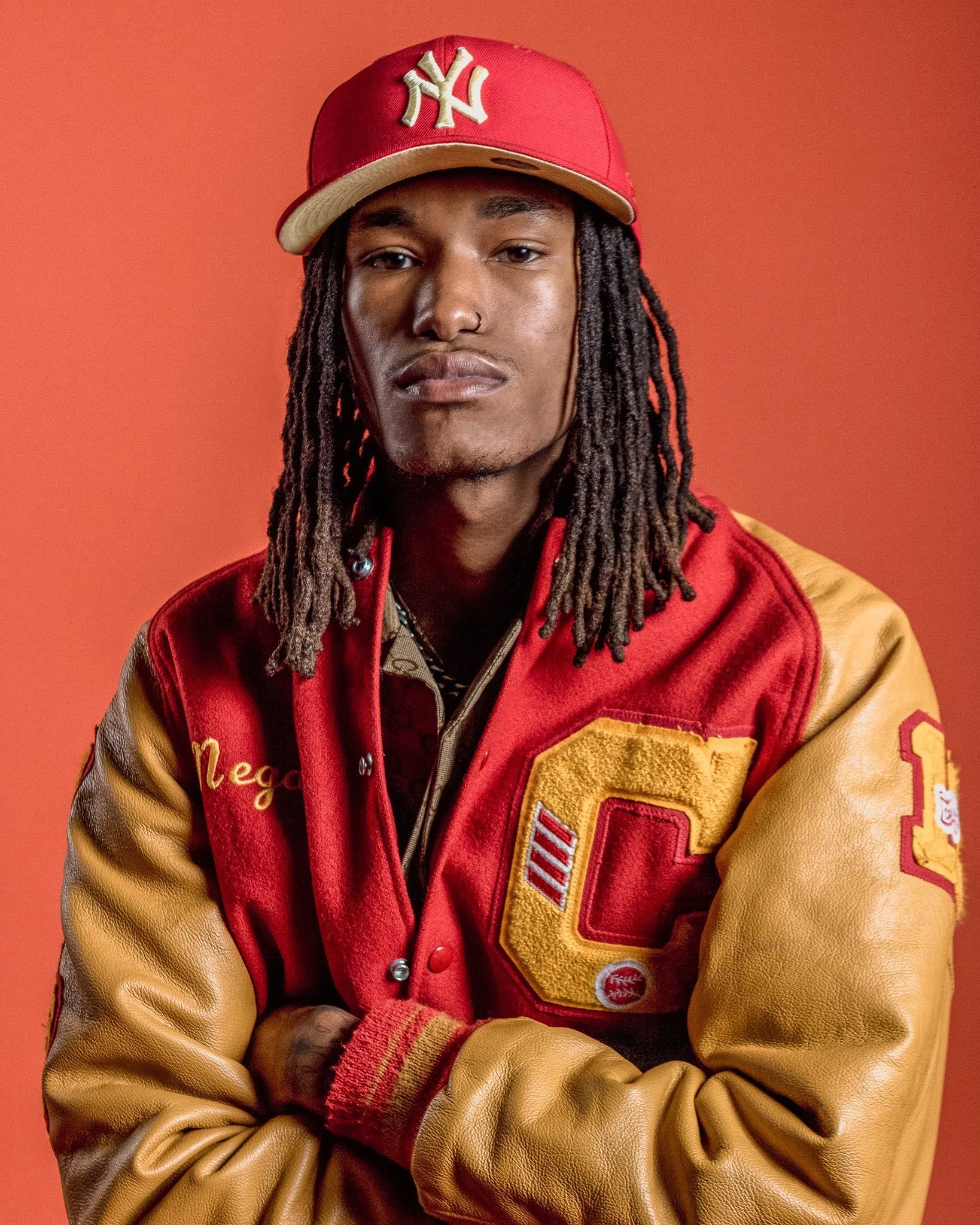 Portrait of a young man with dreadlocks wearing a red and yellow varsity jacket and a red New York Yankees cap against an orange background.