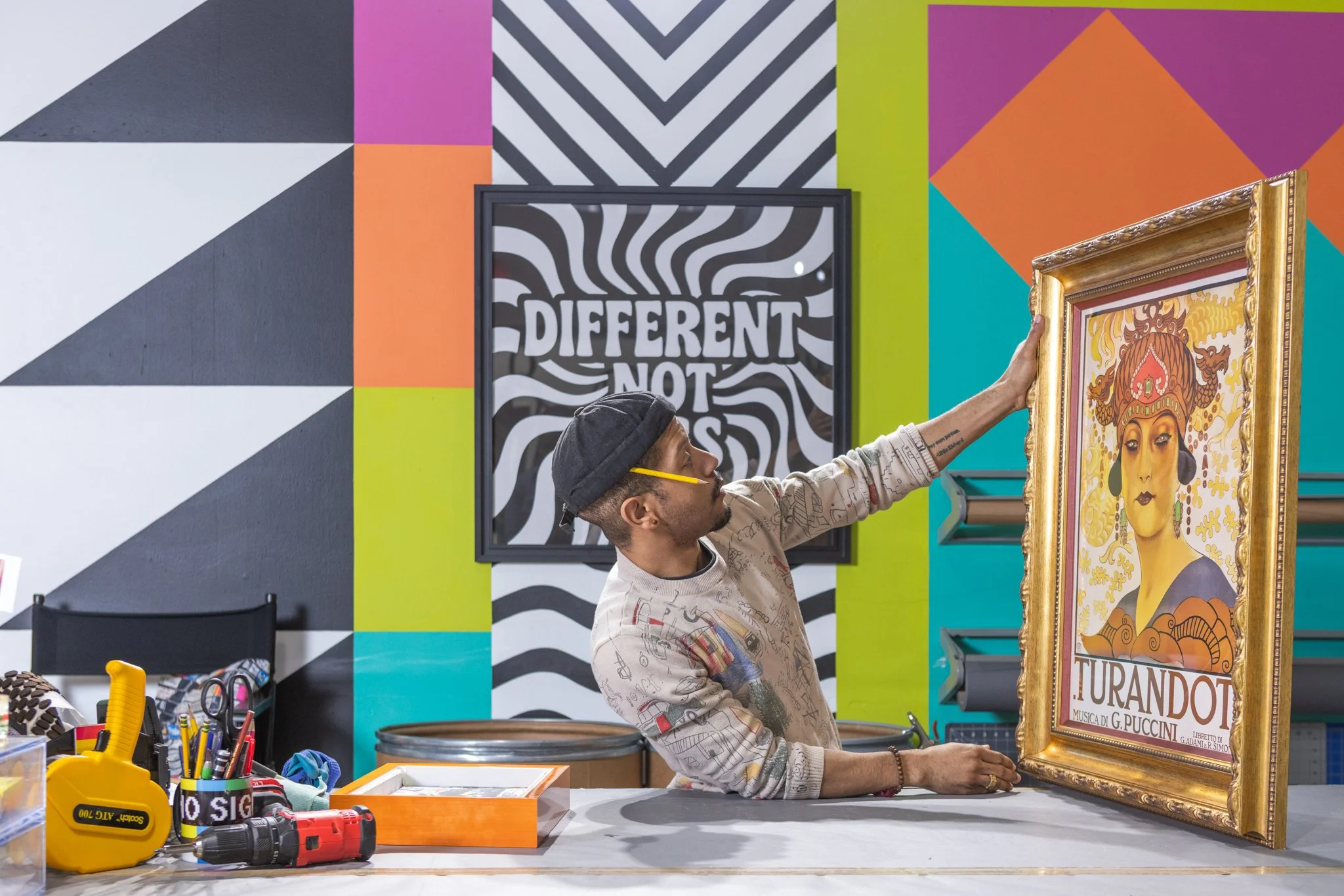 A person in a gray cap and patterned sweatshirt is adjusting or hanging a framed artwork of a woman with a dragon-shaped headdress and ornate gold frame in an art studio with colorful, patterned wall paintings.