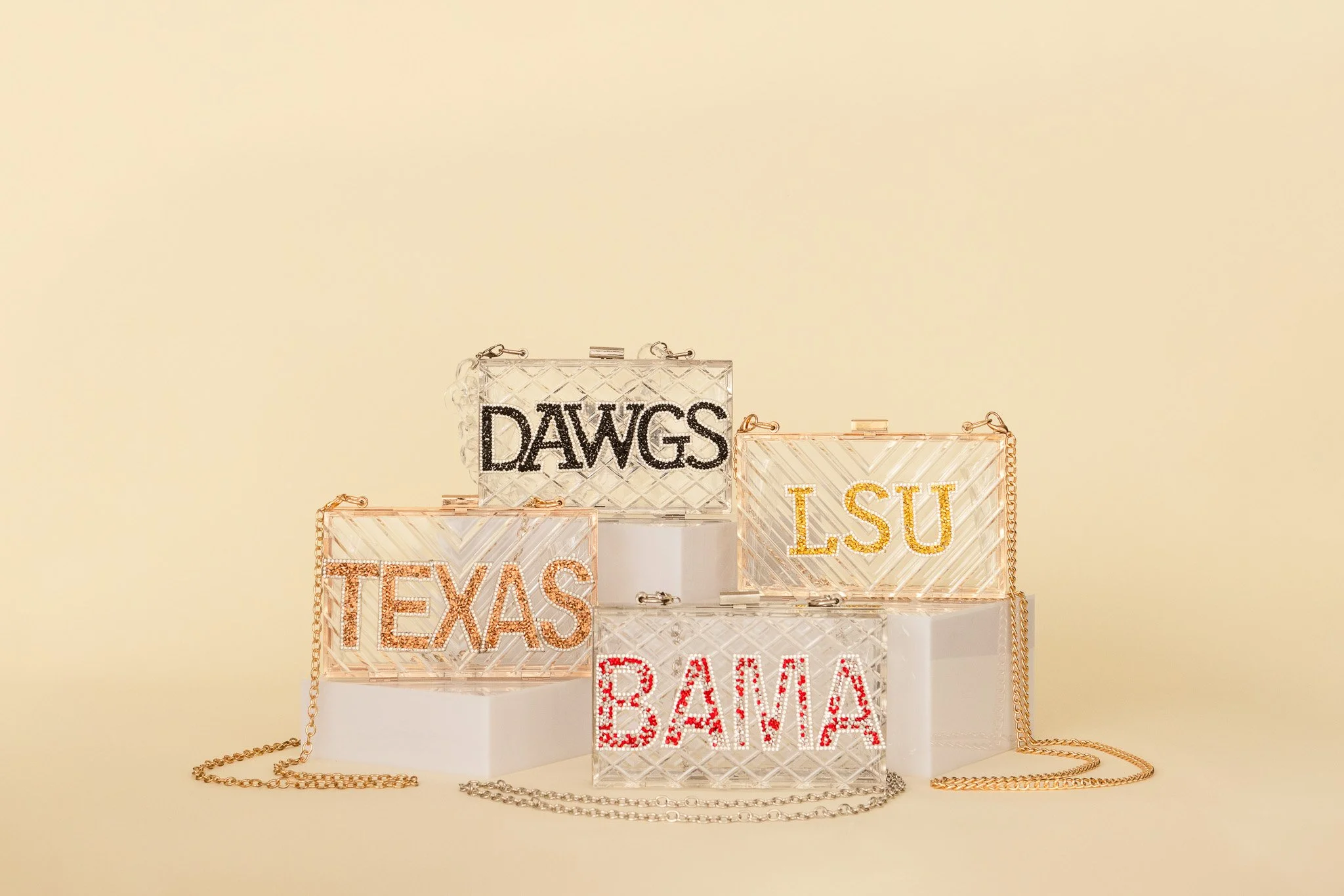 Multiple transparent bags with city names in rhinestones and chains in front of a plain beige background.