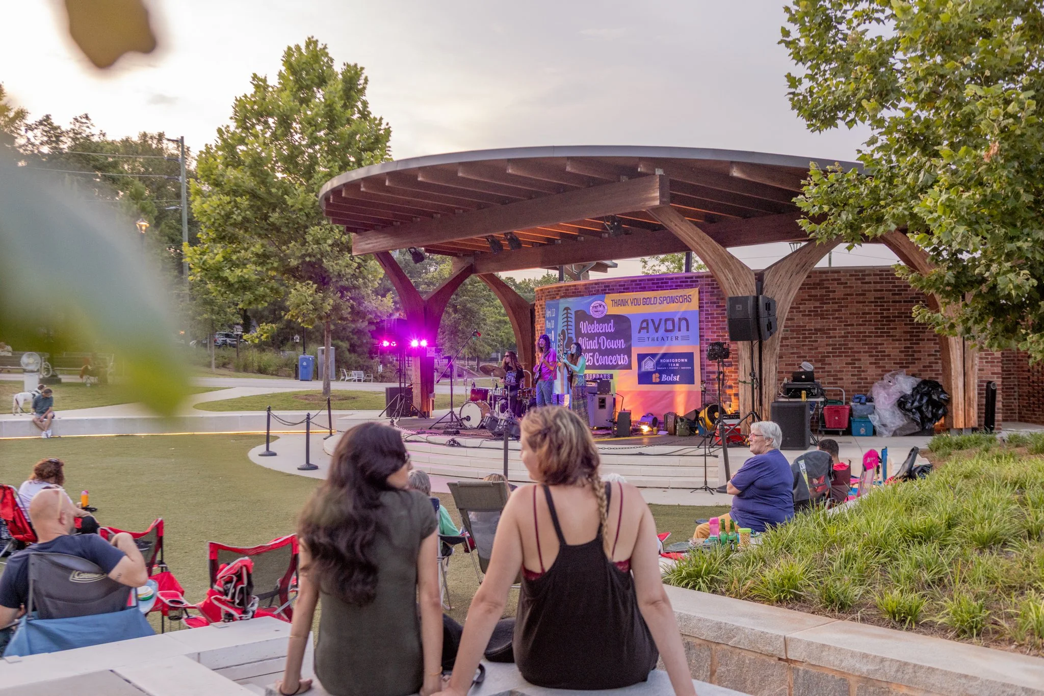 People watching a live band perform on an outdoor stage at sunset in a park, with trees and a grassy area, some audience members sitting on chairs and others on the ground.