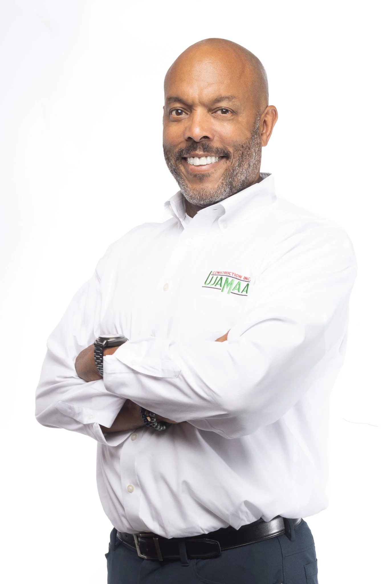 A middle-aged man with a beard and mustache, smiling, wearing a white dress shirt with a logo on the chest, crossed arms, standing against a white background.