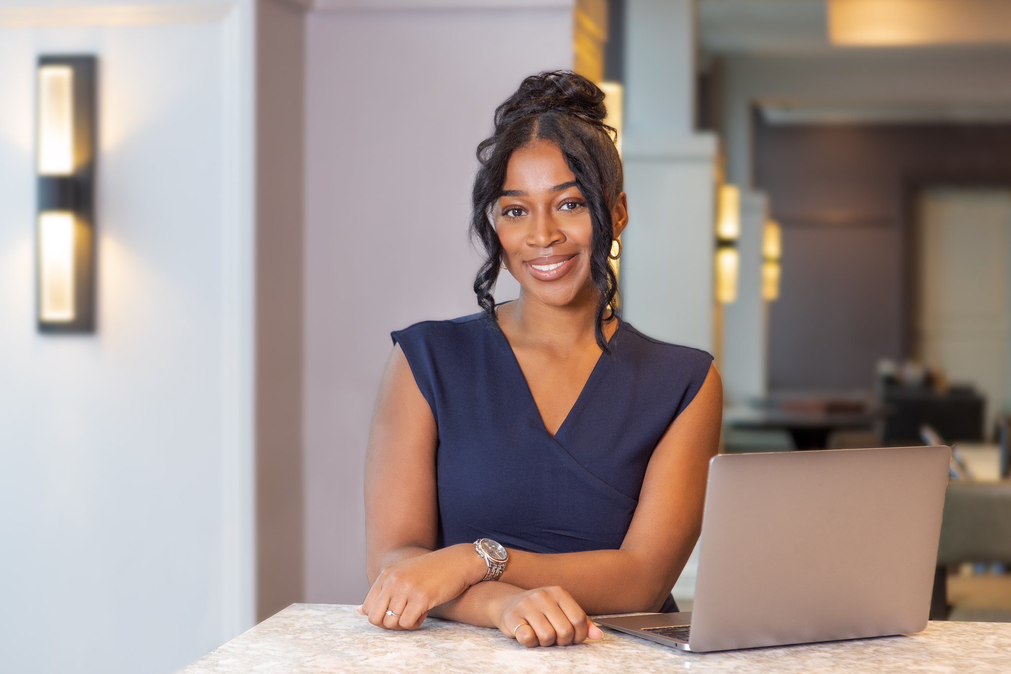 A smiling woman with dark hair styled up and wearing a sleeveless navy dress, sitting at a marble countertop with a laptop in front of her in a well-lit modern interior.