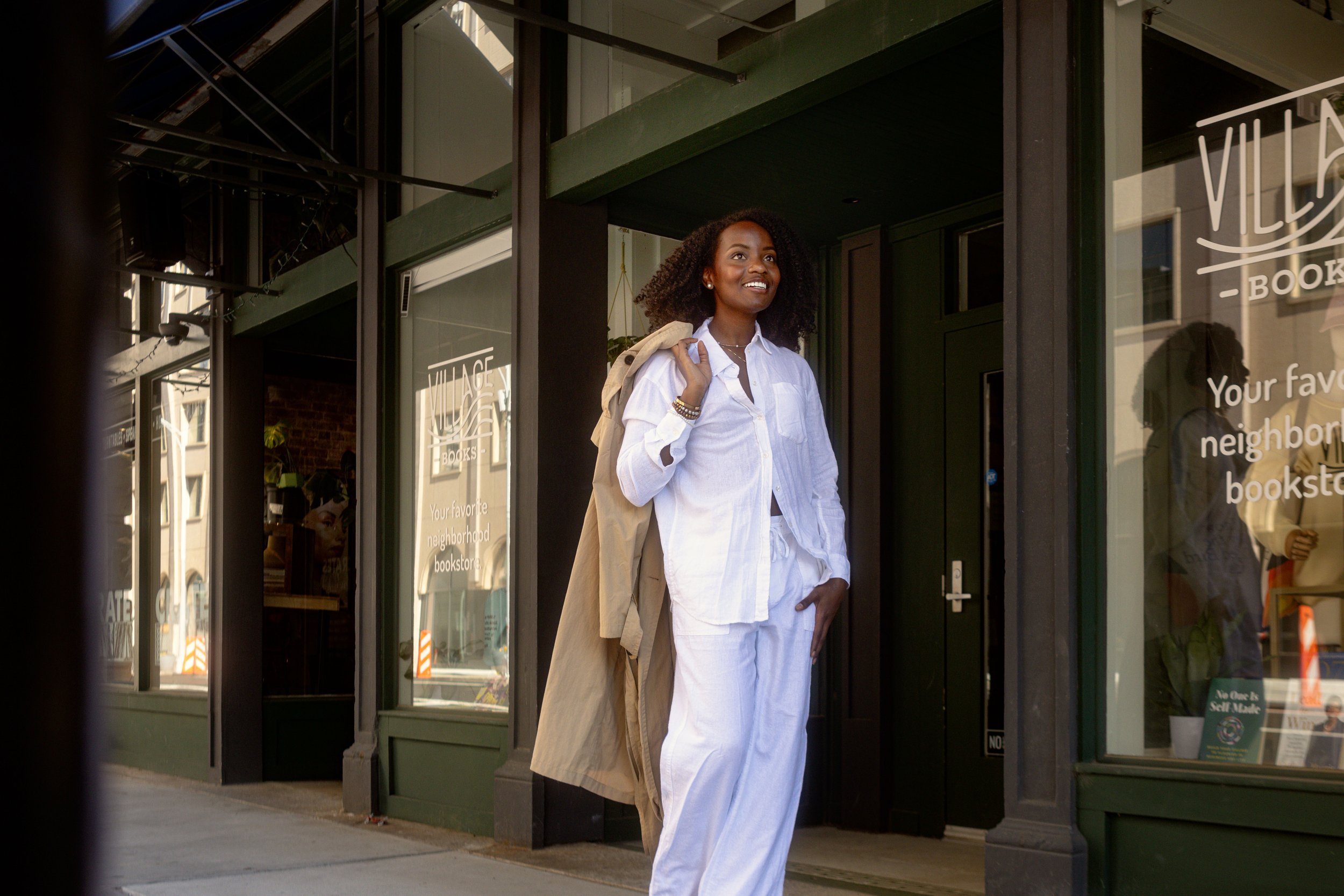 A woman with curly hair wearing a white shirt and white pants, carrying a beige coat over her shoulder, standing outside a bookstore or café storefront with glass windows and green framing, smiling and looking to her left.