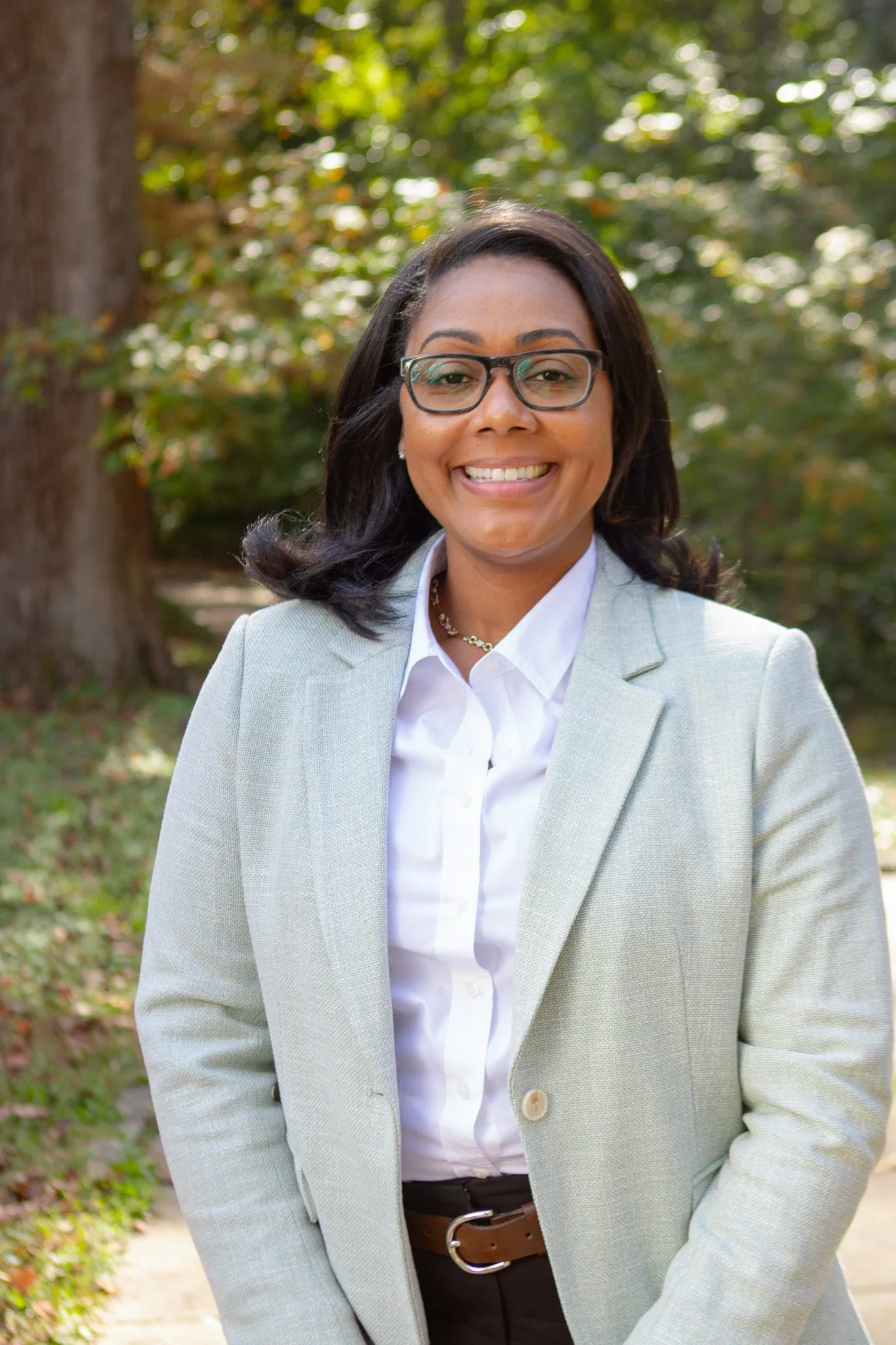 A smiling woman with glasses, wearing a light gray blazer and white shirt, outdoors with green trees and sunlight in the background.