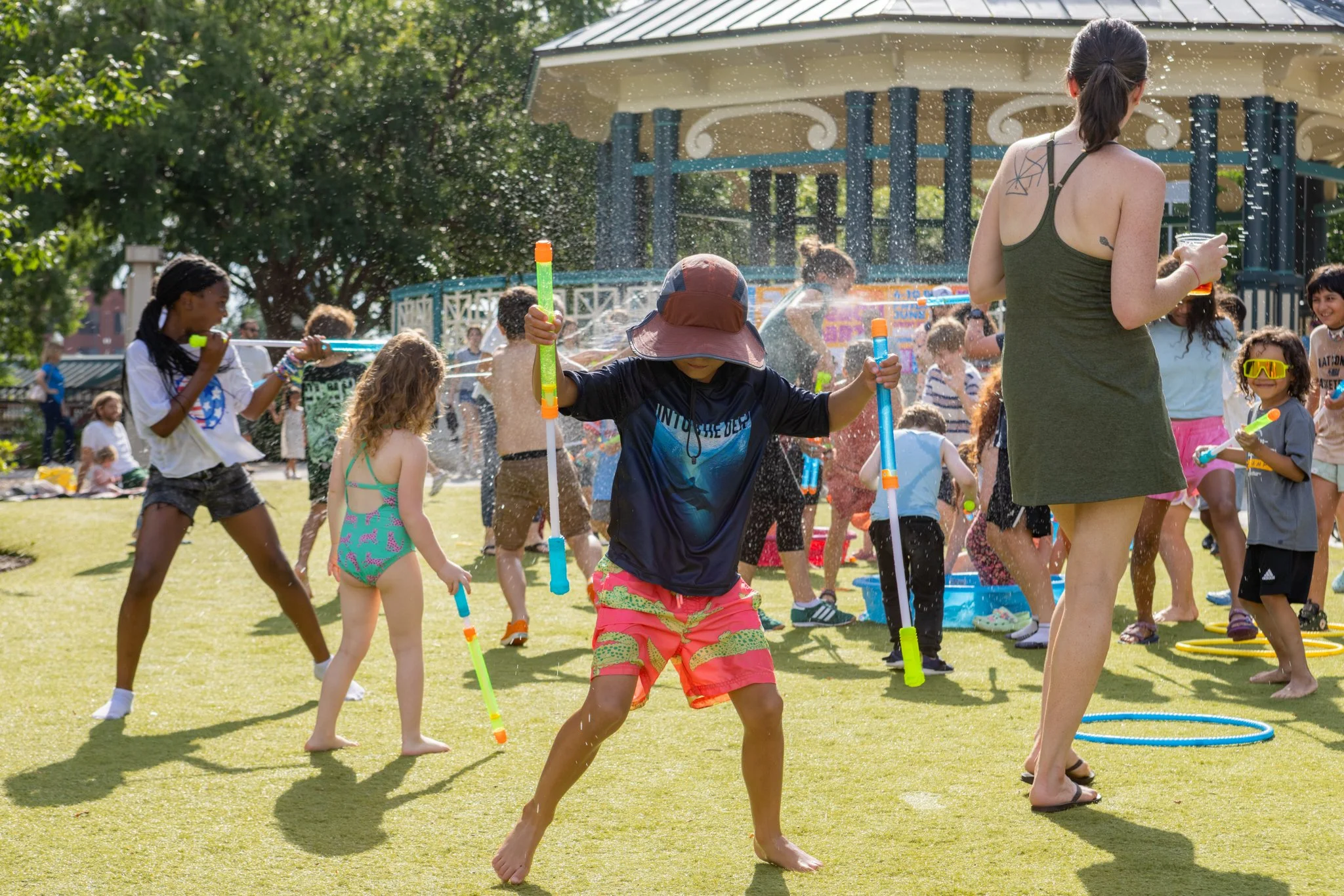 Children and adults playing with water guns outdoors on a sunny day, surrounded by trees and a pavilion.