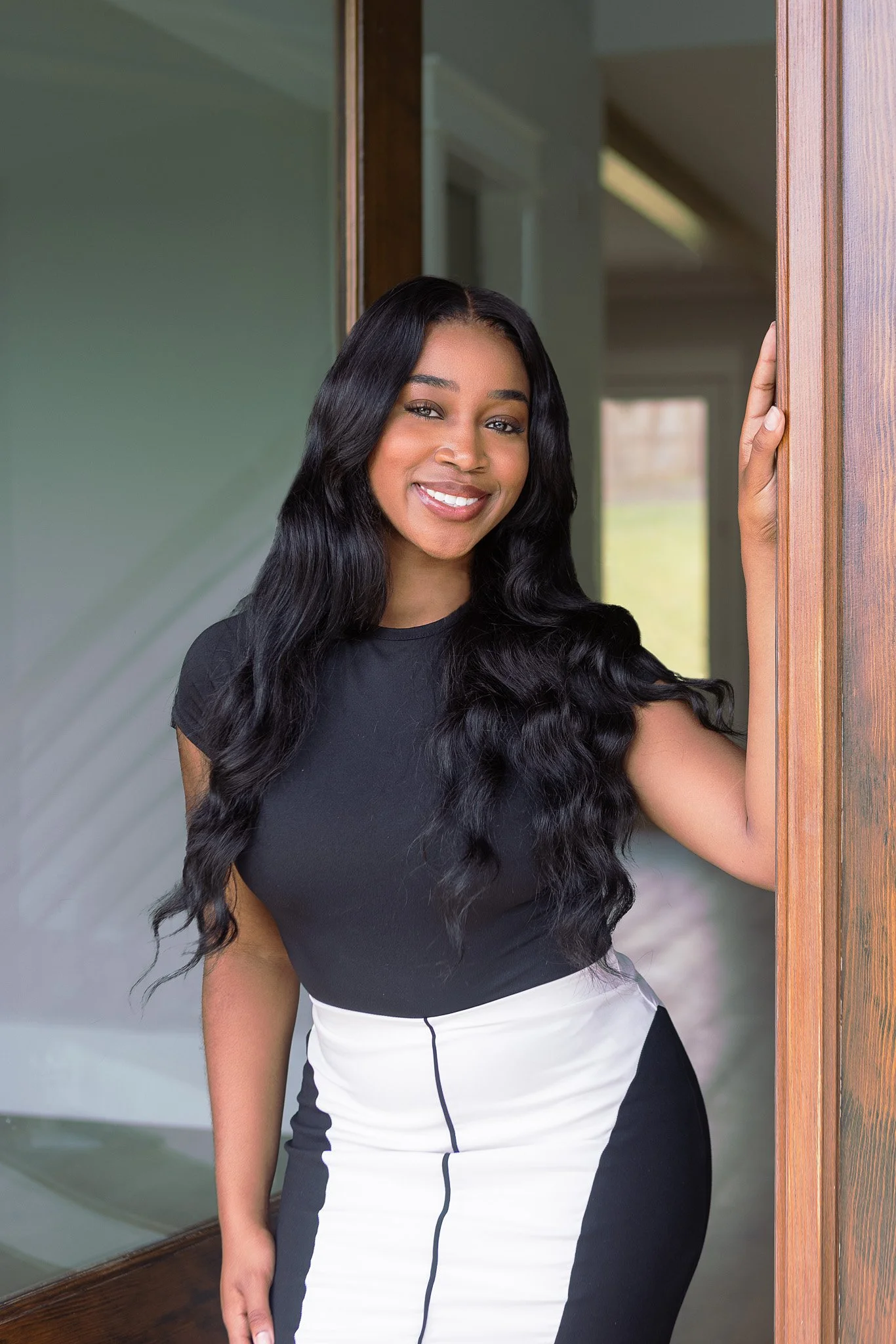 A young woman with long wavy black hair smiling and standing next to a wooden door frame.