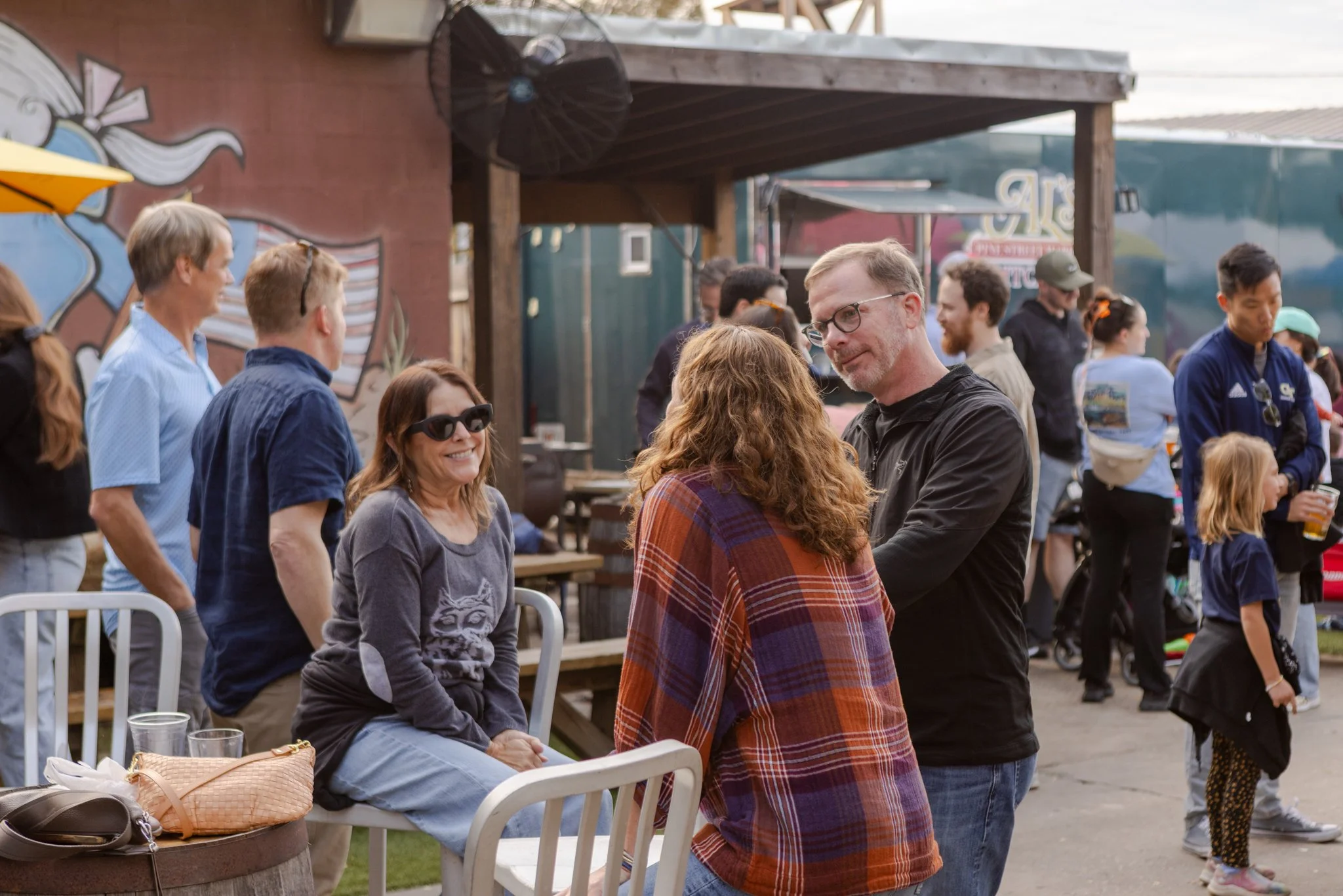 People socializing at an outdoor event, some sitting on chairs and some standing, with a wooden structure and murals in the background.